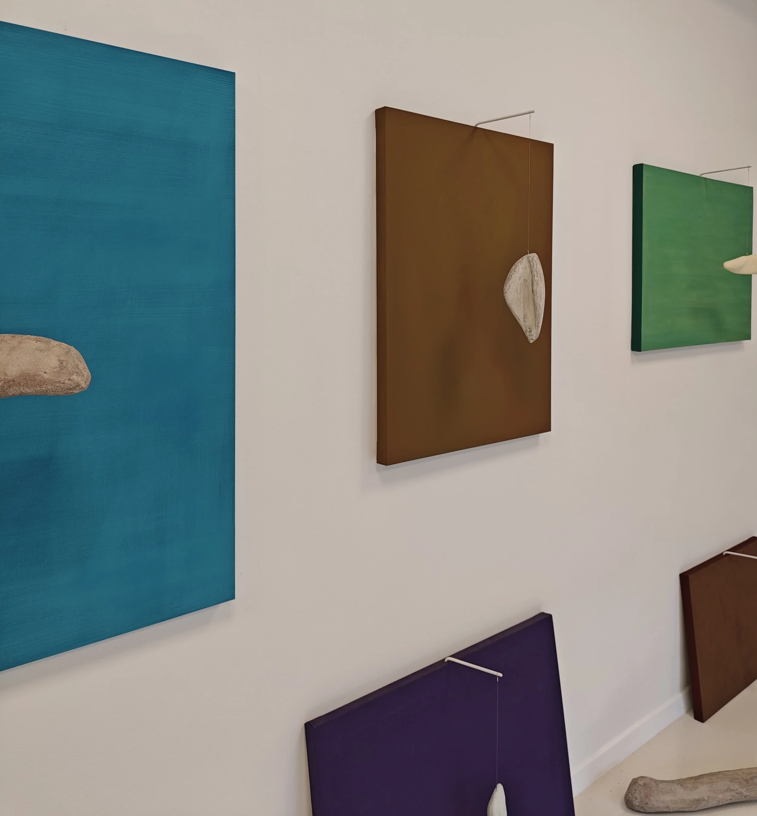 Installazione artistica con dipinti colorati e sculture di pietra su una parete bianca.