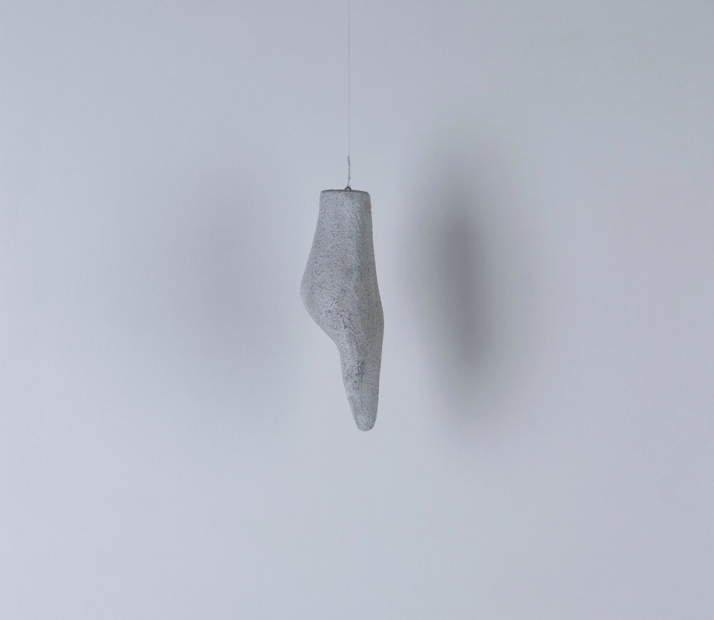 L'installazione artistica di una scultura sospesa a forma di corpo umano, realizzata in materiale grigio con superficie ruvida, appesa al soffitto con un filo.