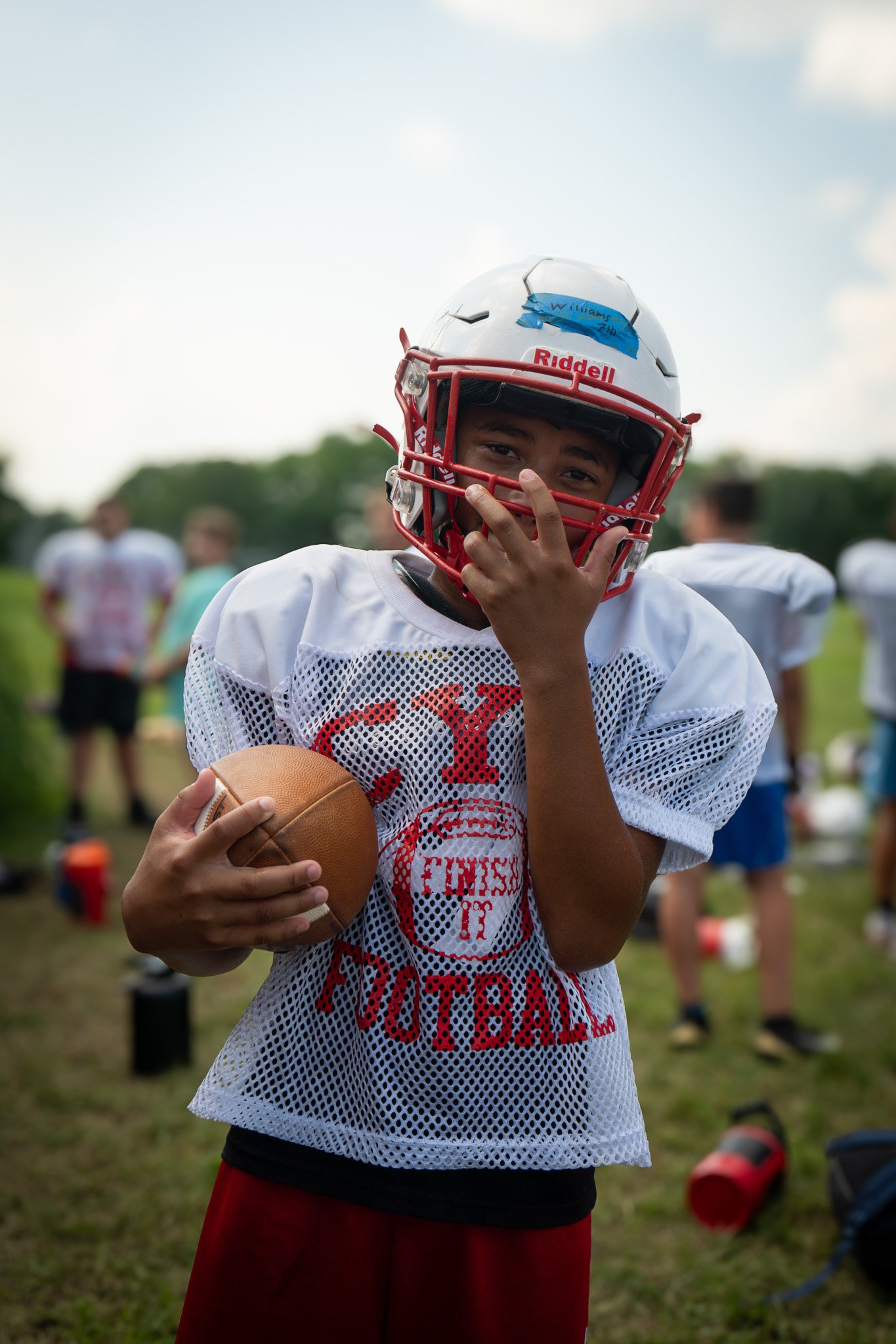 Young boy wearing football gear, holding a football, and smiling outdoors with other children in the background.