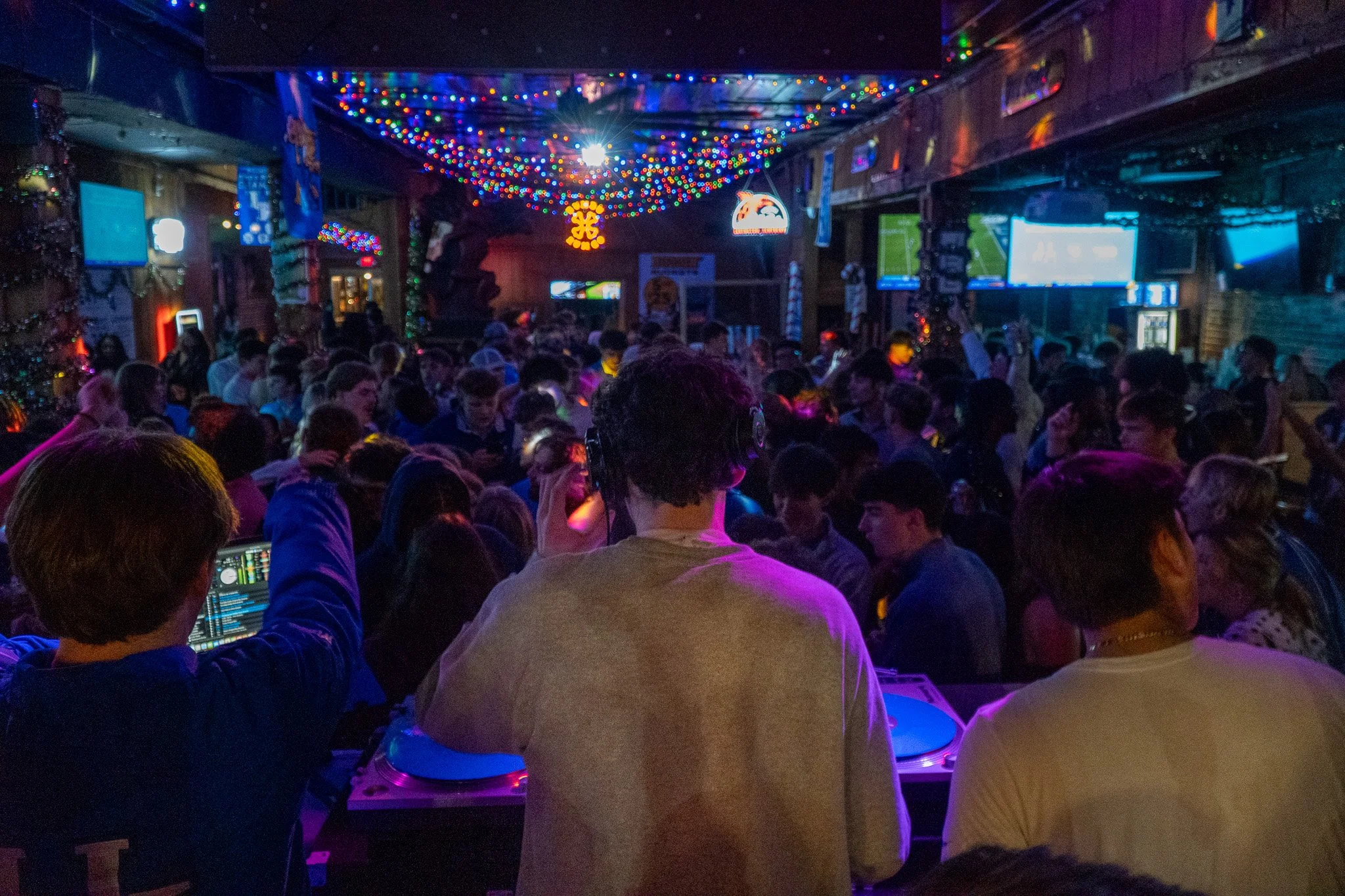 A crowded nightclub with colorful lights, a DJ on the deck, and patrons dancing and socializing.