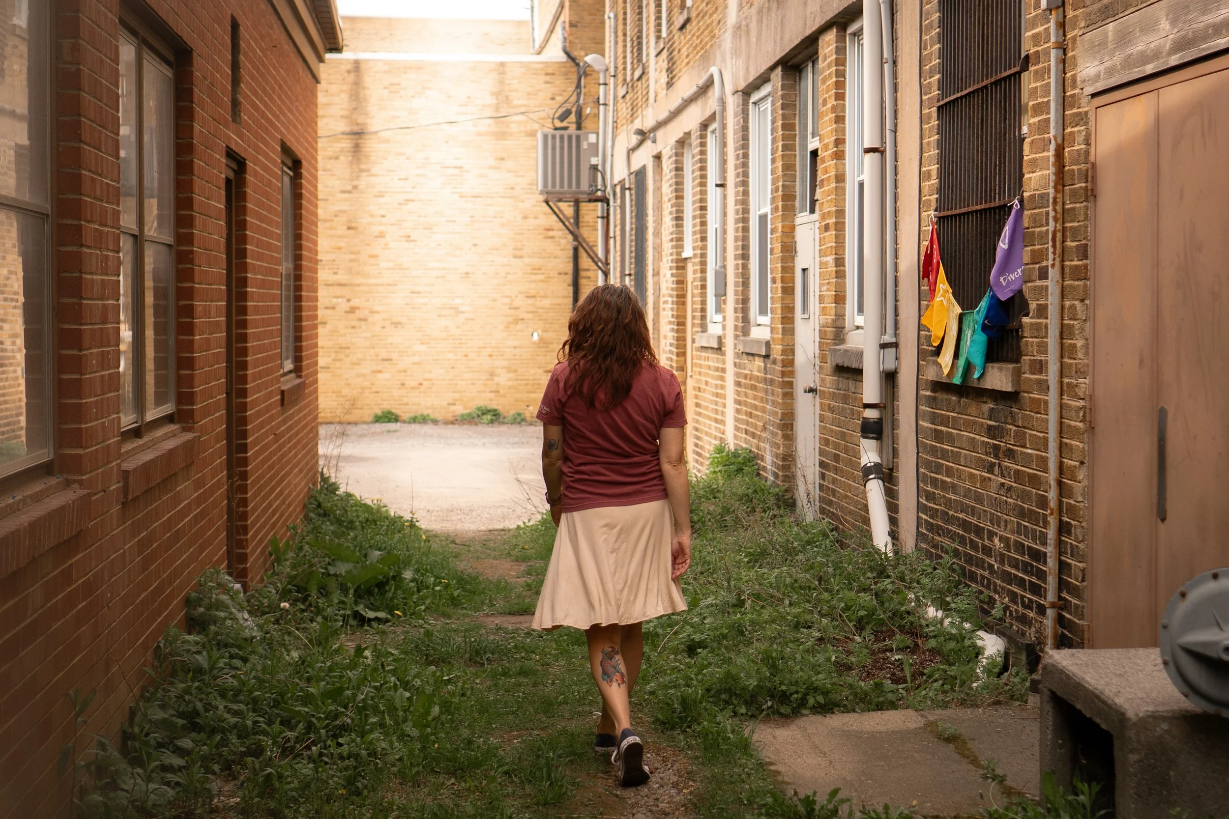 A woman with tattoos on her leg and curly hair walks through a narrow alleyway between brick buildings, with greenery along the ground. She wears a beige skirt and a maroon shirt, and is photographed from behind.