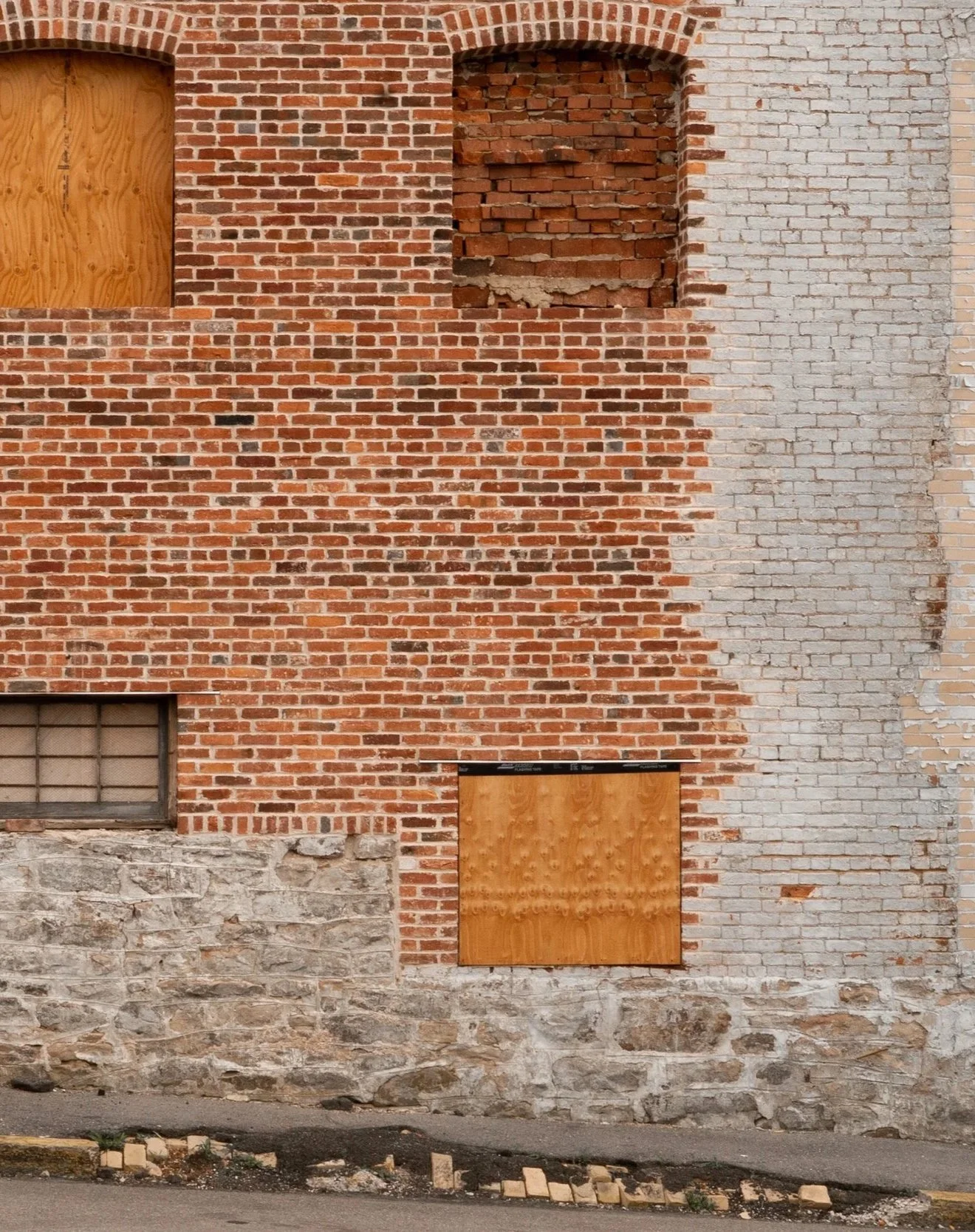 A brick building facade with boarded-up windows and a small section of stone wall. The bricks are varying shades of red and some areas have exposed brickwork and gaps.