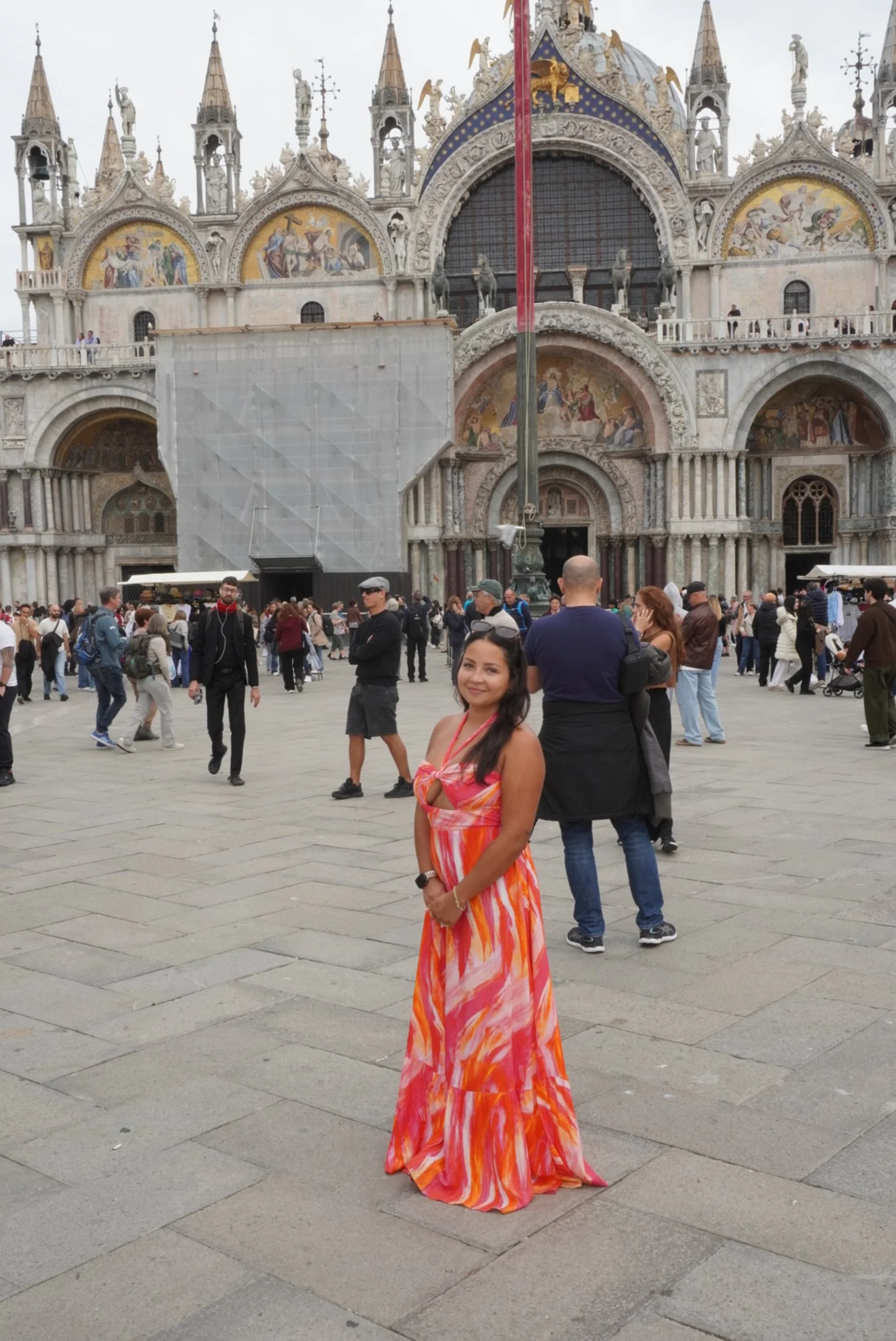 A woman in a colorful orange and pink dress standing in front of St. Mark's Basilica in Venice, Italy, with many tourists in the background.