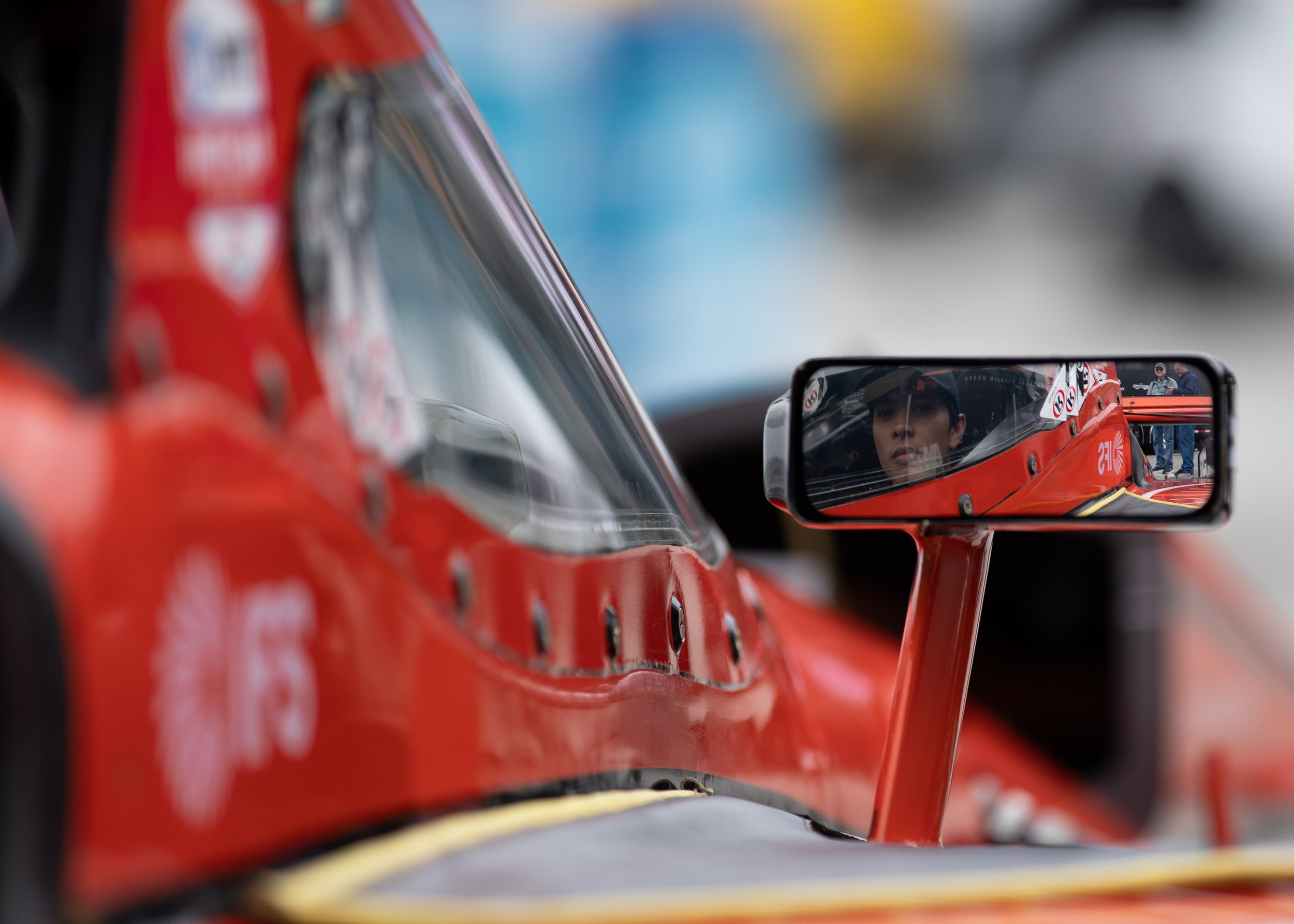 Close-up of a red race car with a side mirror showing the face of a driver wearing a helmet, and a blurred background with people.