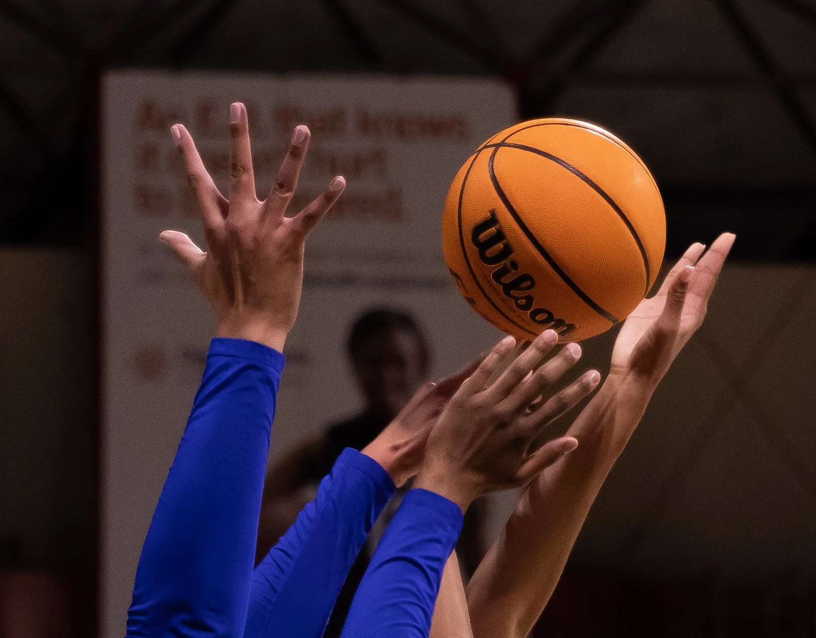 Close-up of multiple hands reaching for a Wilson basketball during a game or practice, with players wearing blue jerseys, against a blurred indoor gym background.