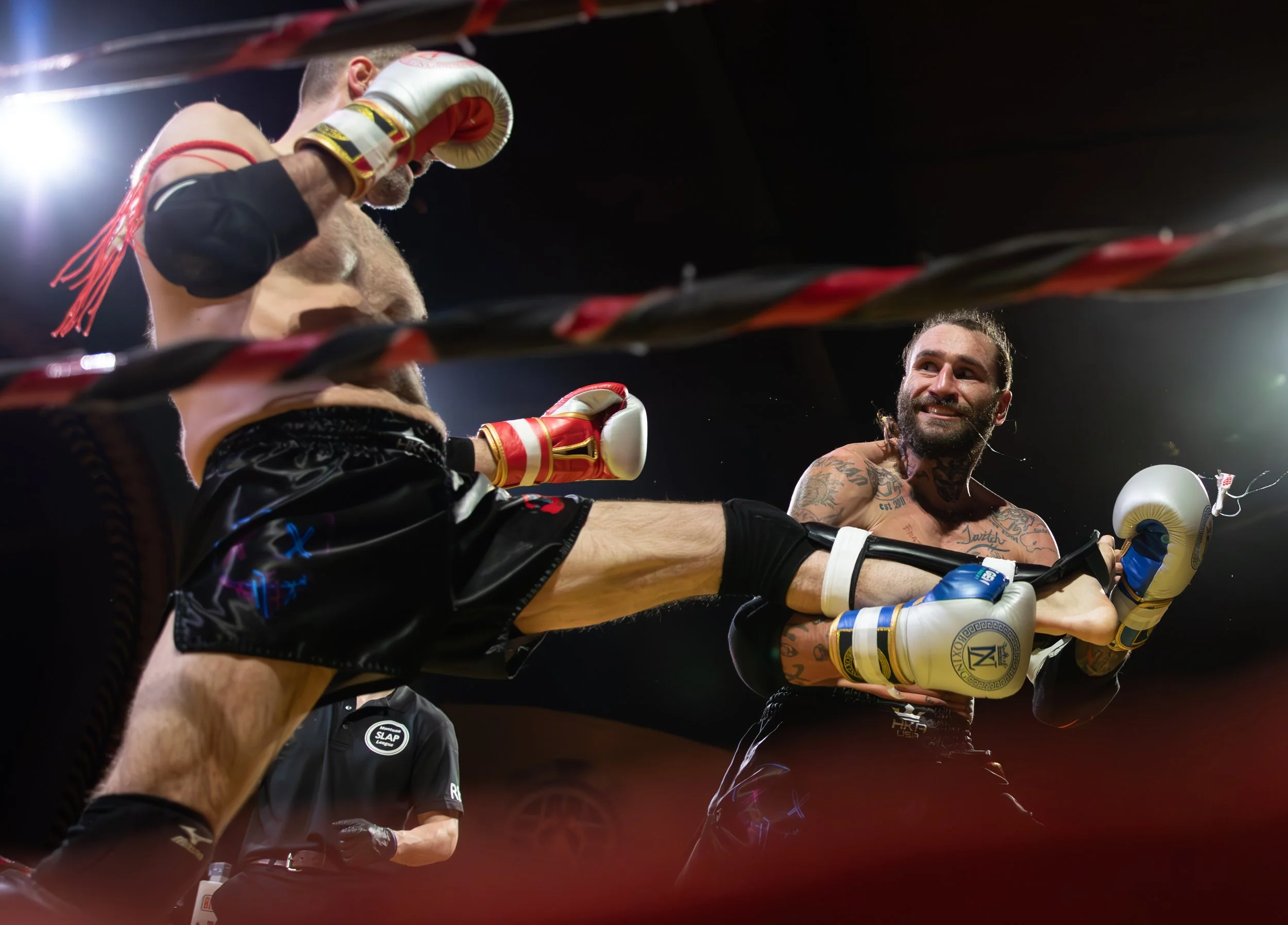 Two shirtless male boxers with tattoos exchanging punches inside a boxing ring, with one delivering a kick to the other's midsection, under bright overhead lights and a dark background.