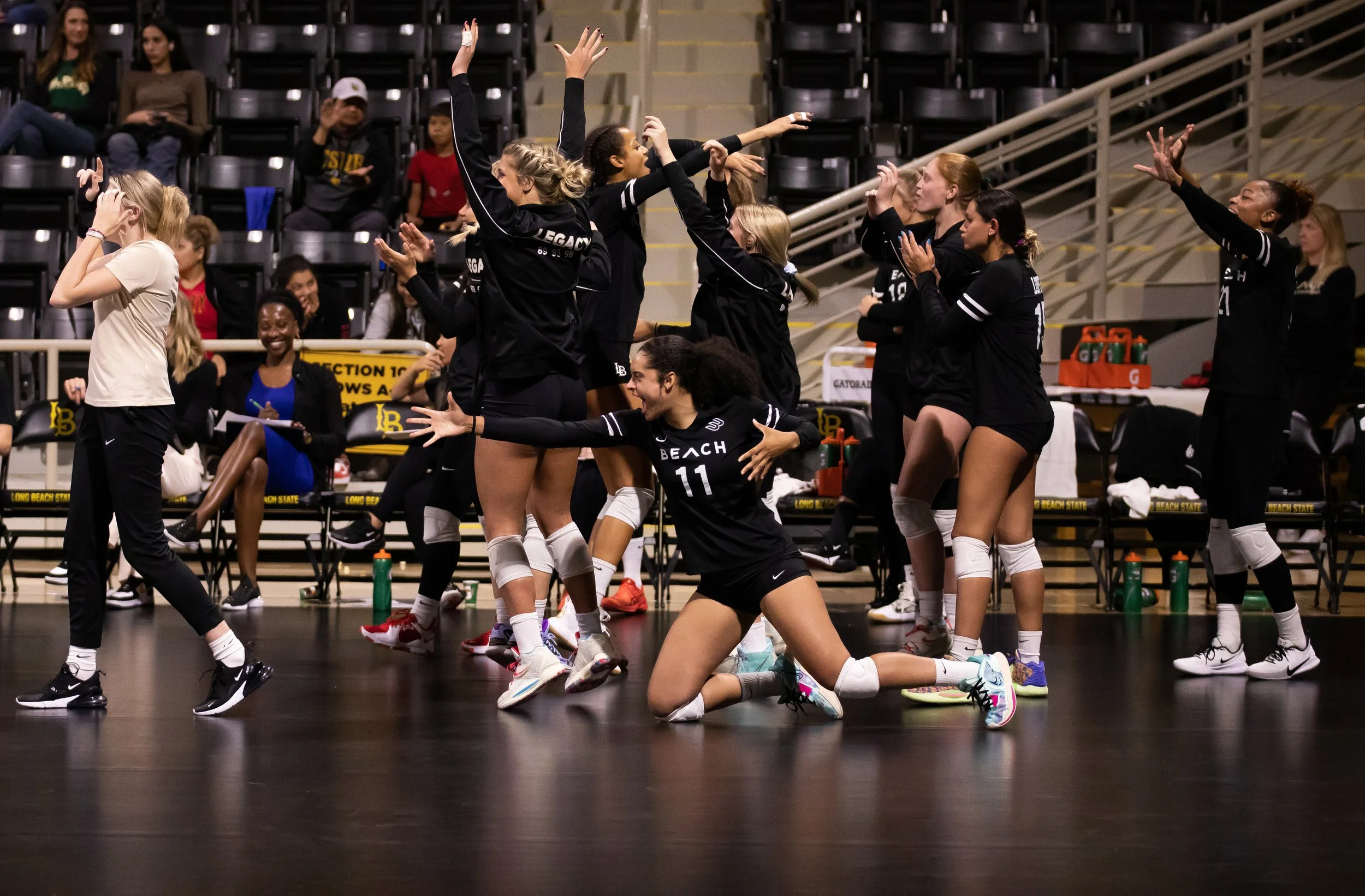 Group of female volleyball players celebrating on the court during a match, some are jumping, others are kneeling or clapping, with spectators in the background.