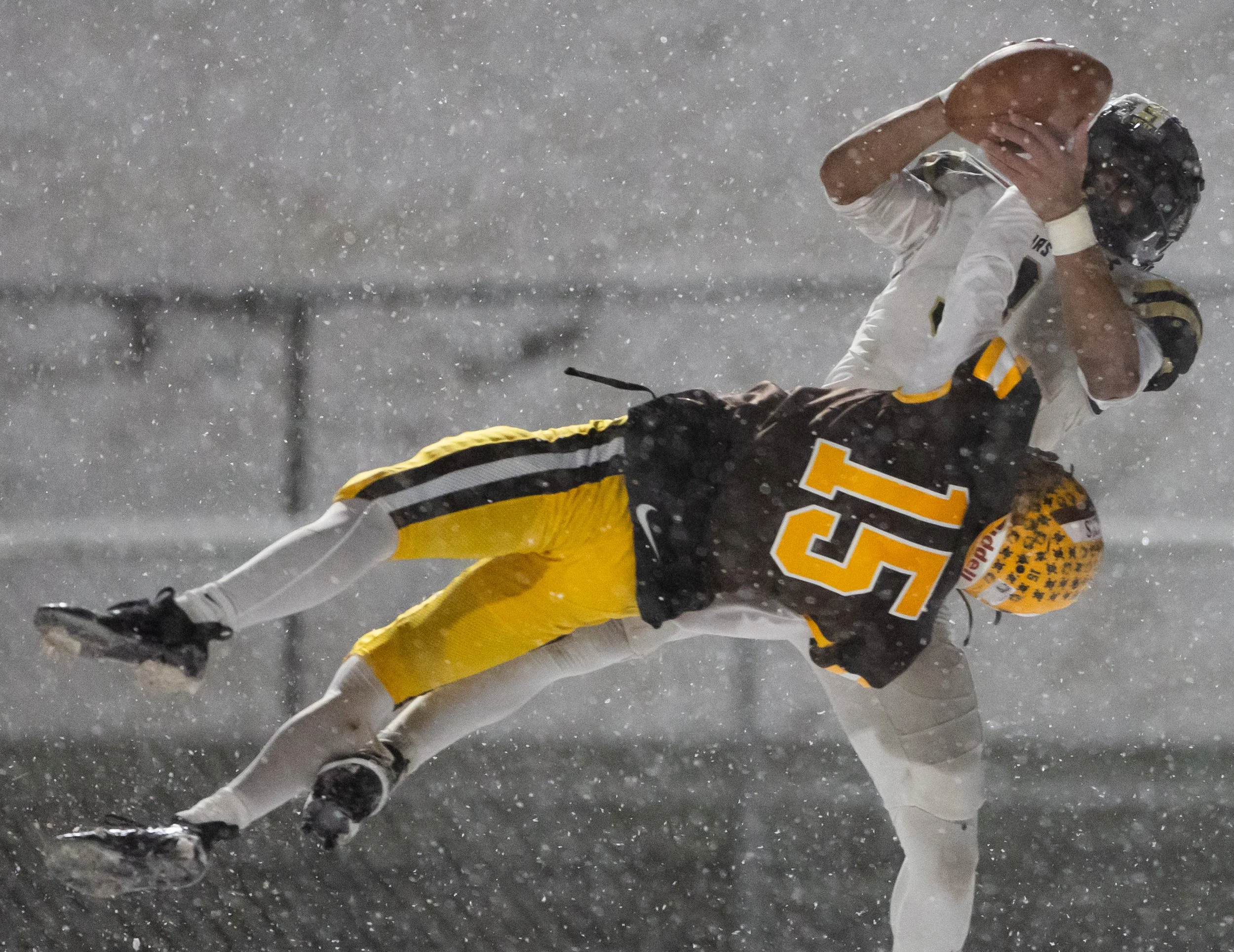 Two football players in rainstorm, one in white uniform attempting to catch the ball, the other in black and yellow uniform falling.