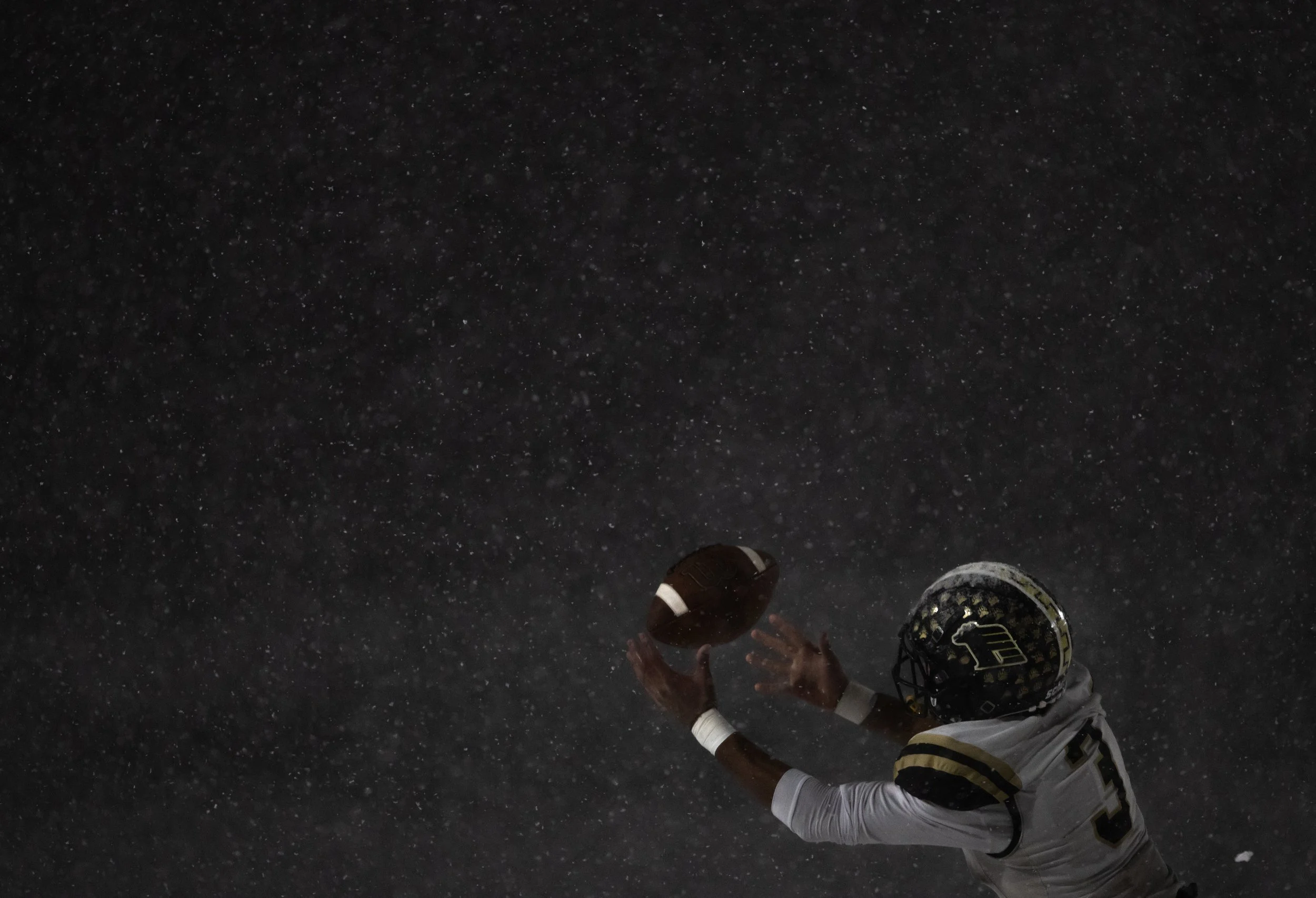 A football player wearing a helmet and white jersey reaching out to catch a football during a snowstorm at night.