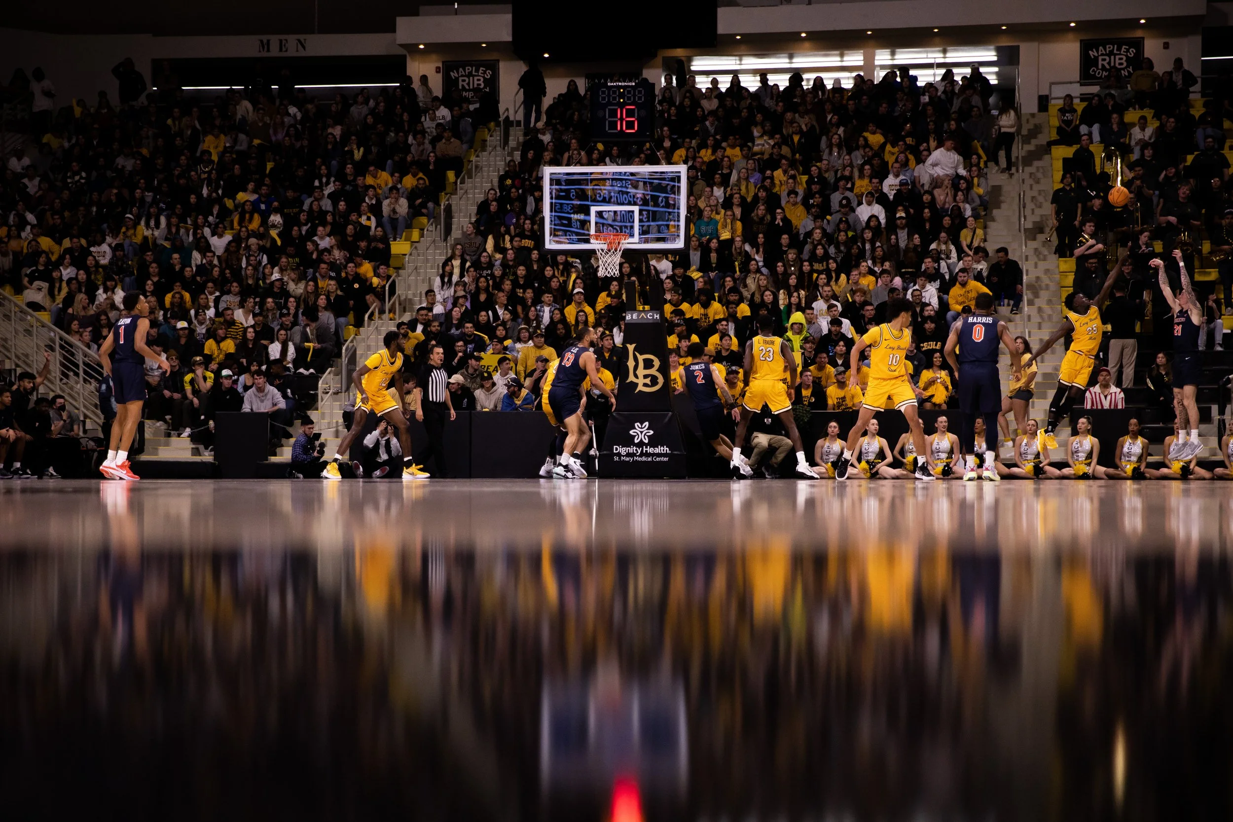 Indoor basketball game with players, cheerleaders, and a large crowd in the stands.