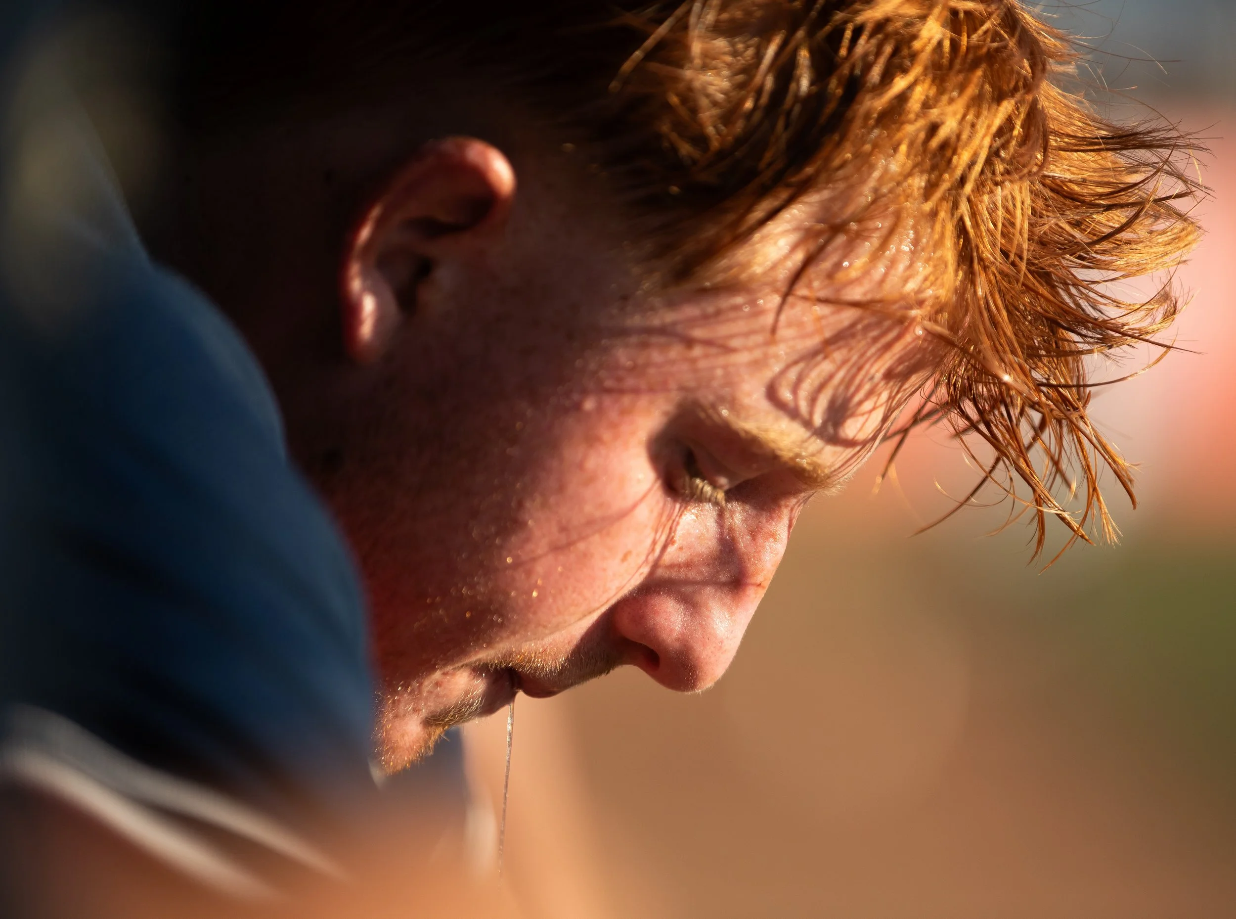 A close-up side profile of a young man with wet, reddish hair, looking down with sunlight illuminating his face.