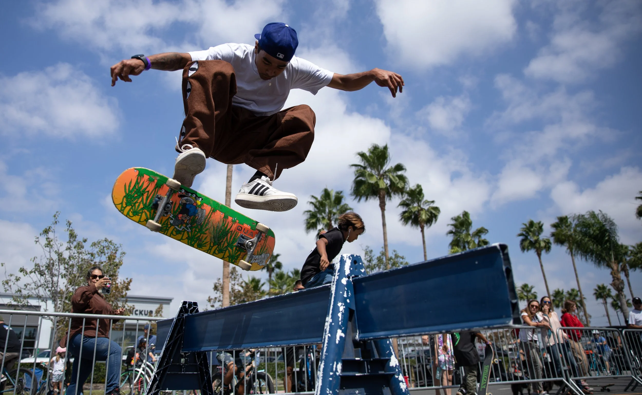 Skateboarder performing a jump over a blue rail at an outdoor skate park, with onlookers and palm trees in the background under a partly cloudy sky.