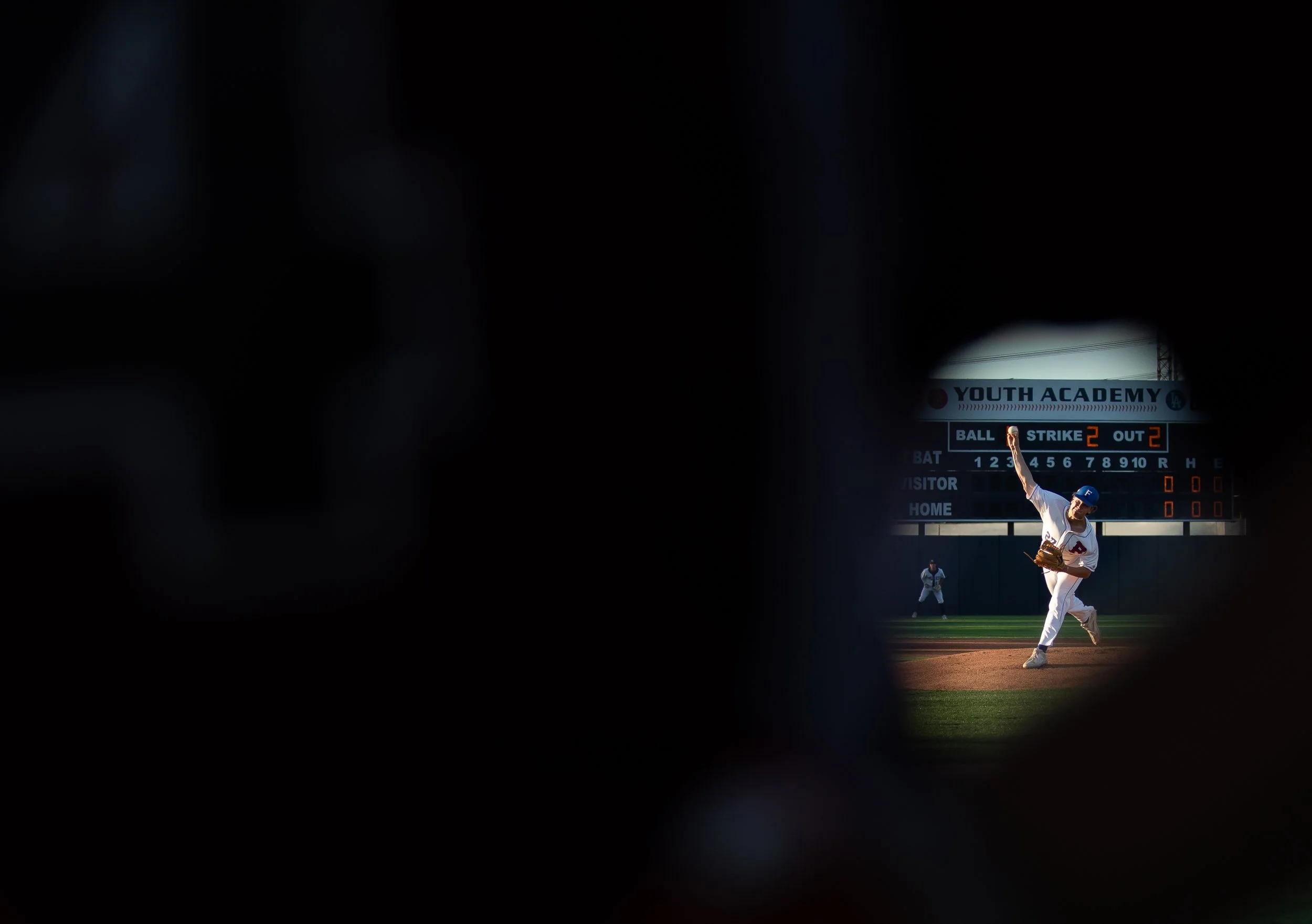 A baseball game viewed through a small gap, with a player in a white uniform catching a ball on the field under a scoreboard that reads "YOUTH ACADEMY" in the background.