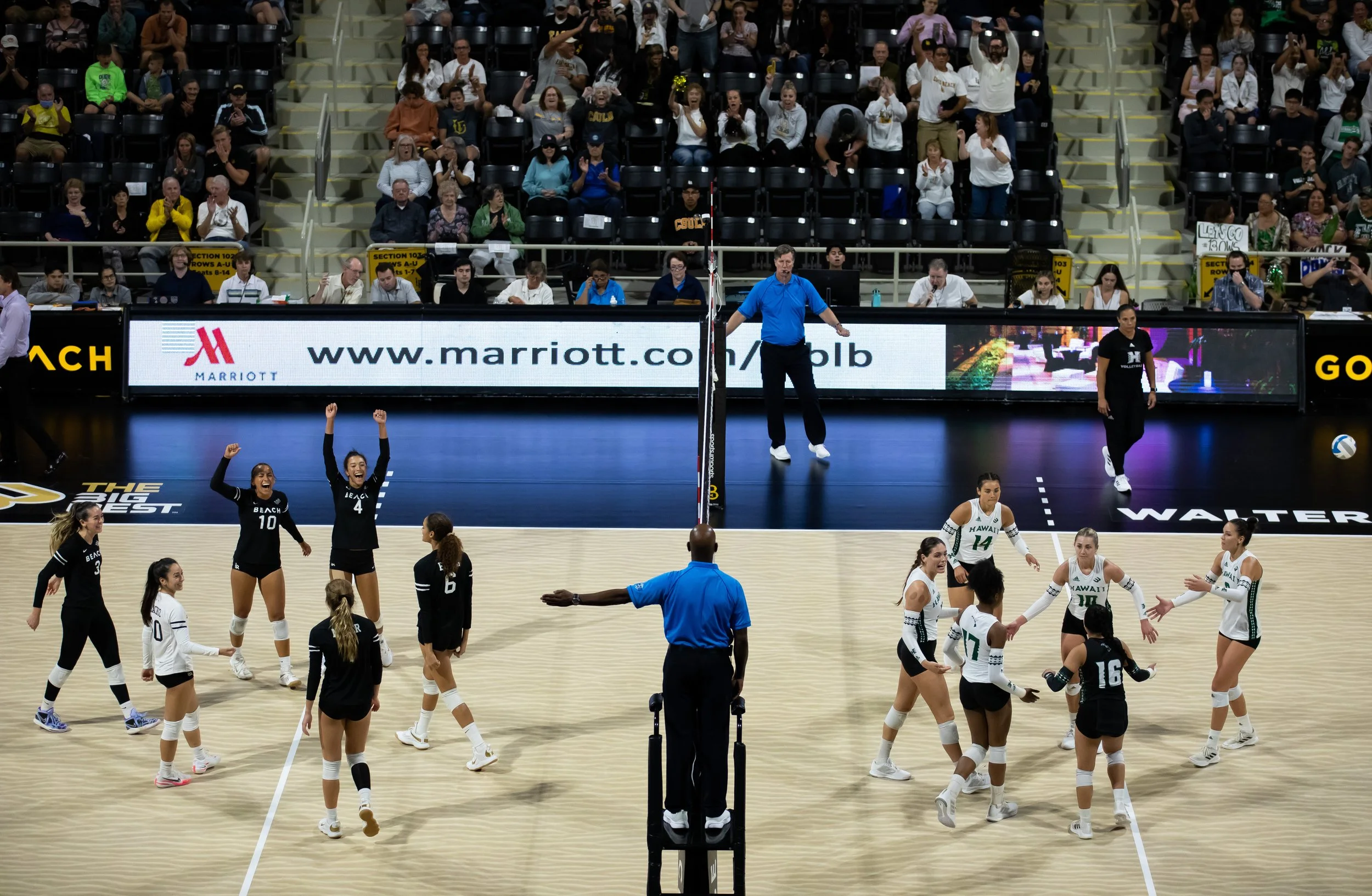 Two women's volleyball teams on an indoor court, celebrating before a match, with a crowd seated in the background, a referee in the foreground, and digital advertisements along the sidelines.