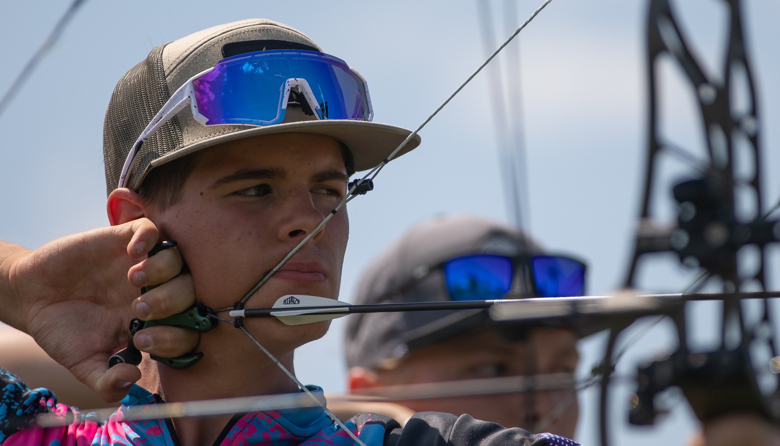 A young male archer wearing sunglasses and a cap, drawing a bow and aiming an arrow, with another person partially visible behind him.