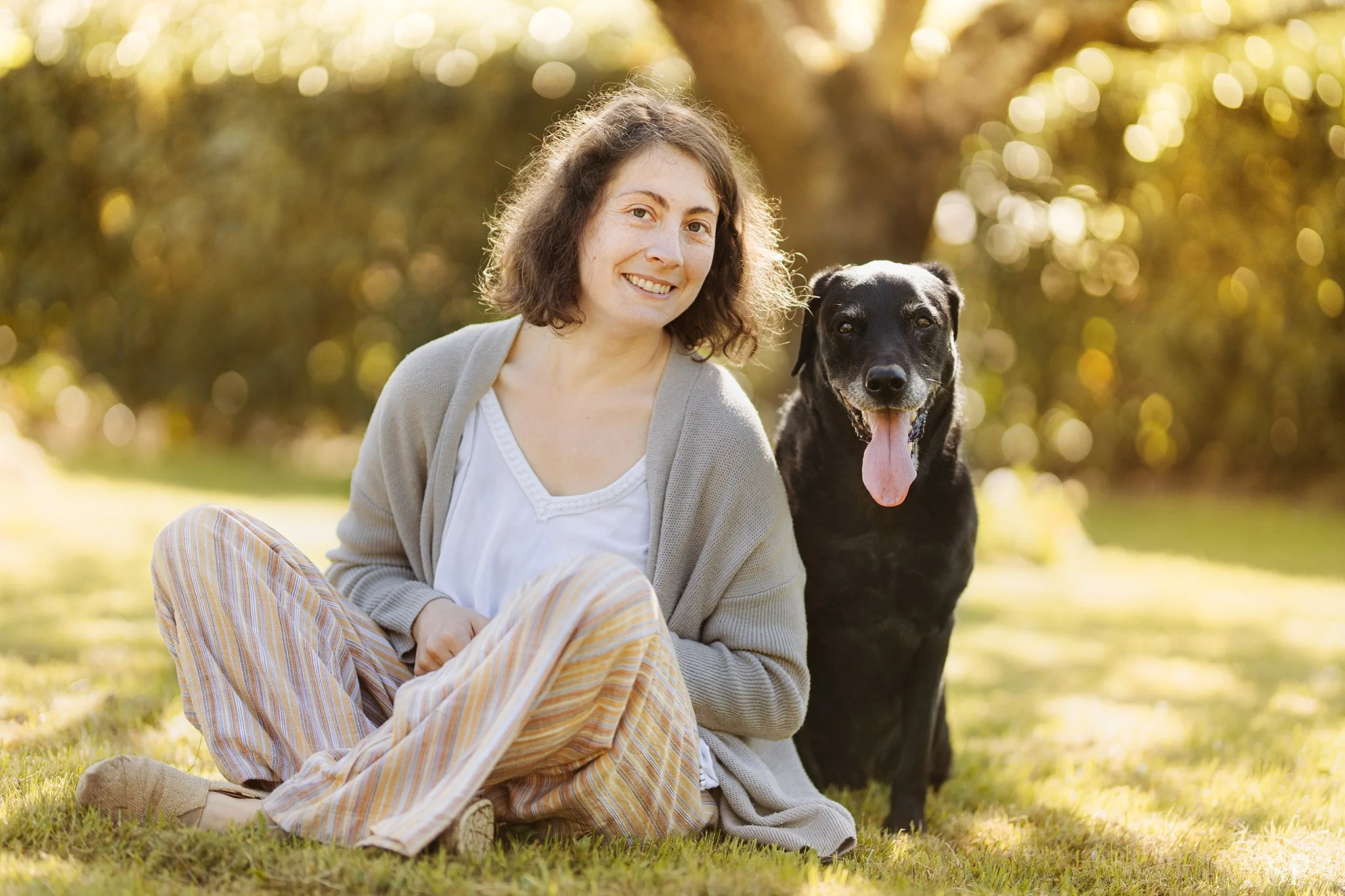 Une femme souriante assise sur l'herbe, à côté d'un chien noir qui a la langue sortie, dans un parc ensoleillé avec des arbres en arrière-plan.