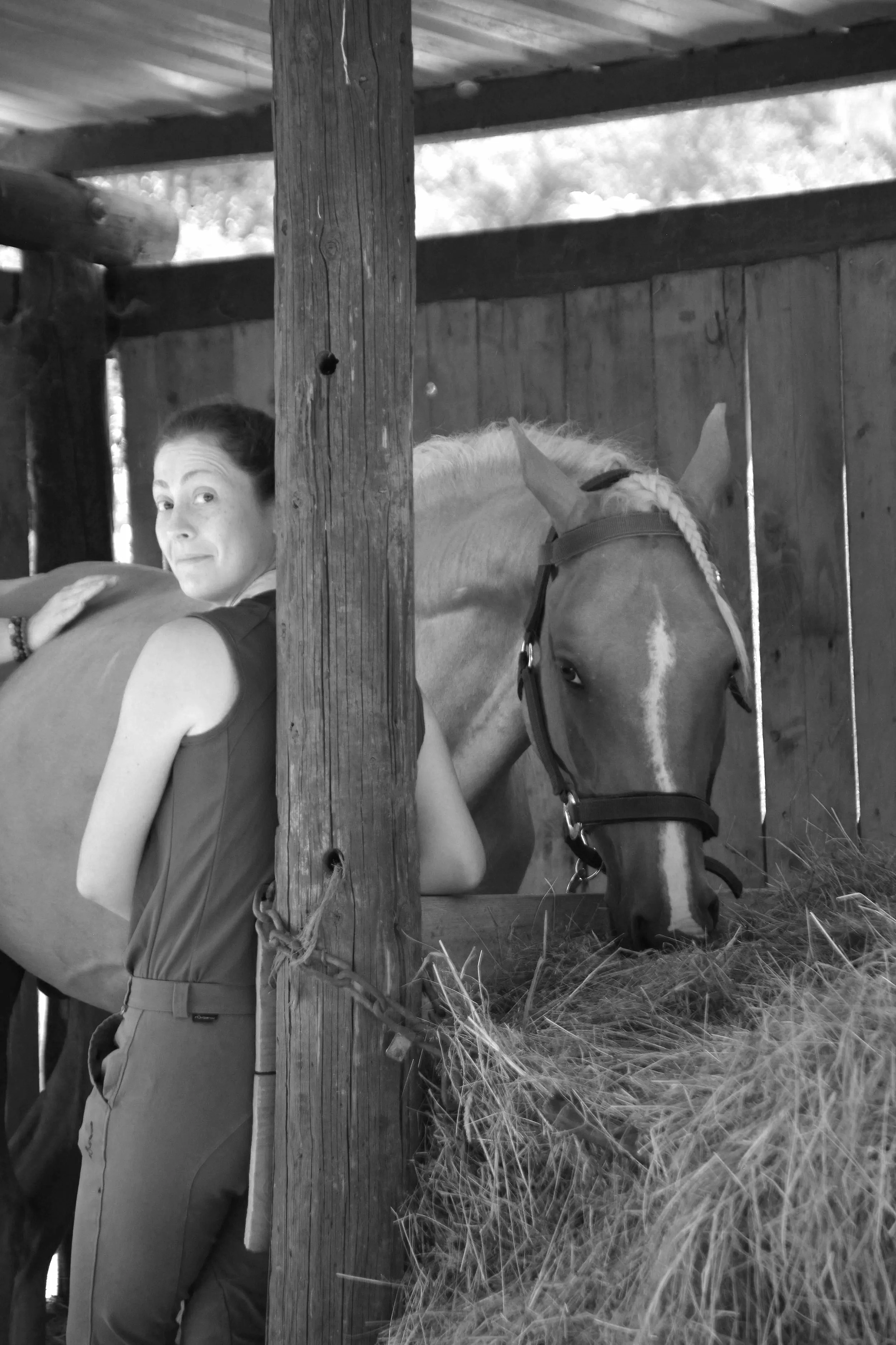 Une femme et un cheval dans un box en bois, la femme regarde vers l'appareil photo et le cheval mange de la paille.