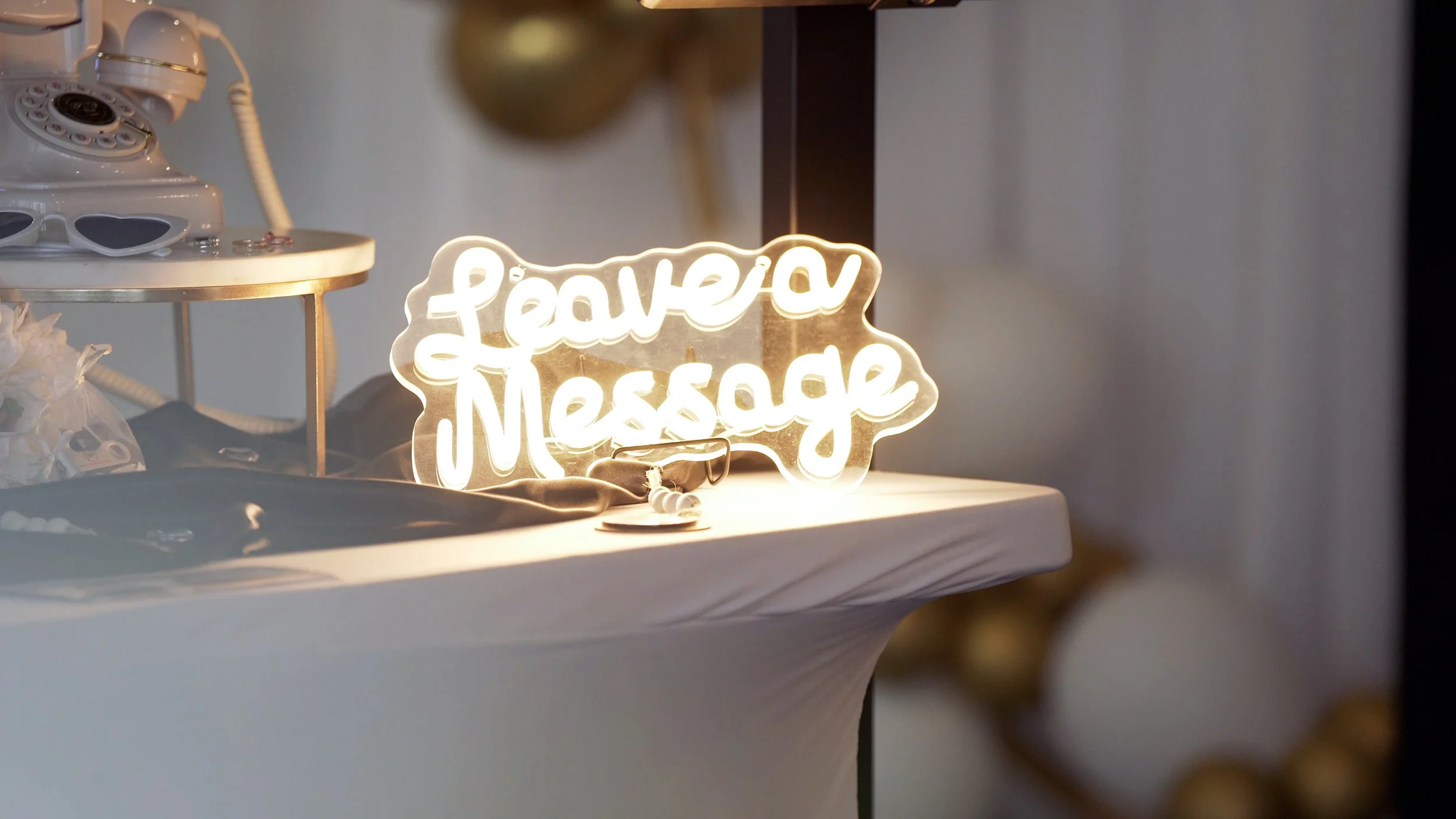 Neon sign that reads 'Leave a Message' placed on a table with miscellaneous items, including a small container and a cloth. Audio Guestbook