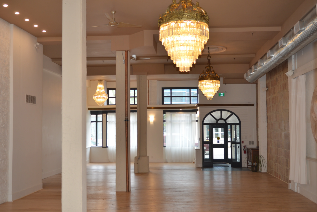 Interior of a spacious room with wooden floors, white walls, several chandeliers, large windows with sheer curtains, an arched glass door, and a brick accent wall. The Avenue Room Saskatoon