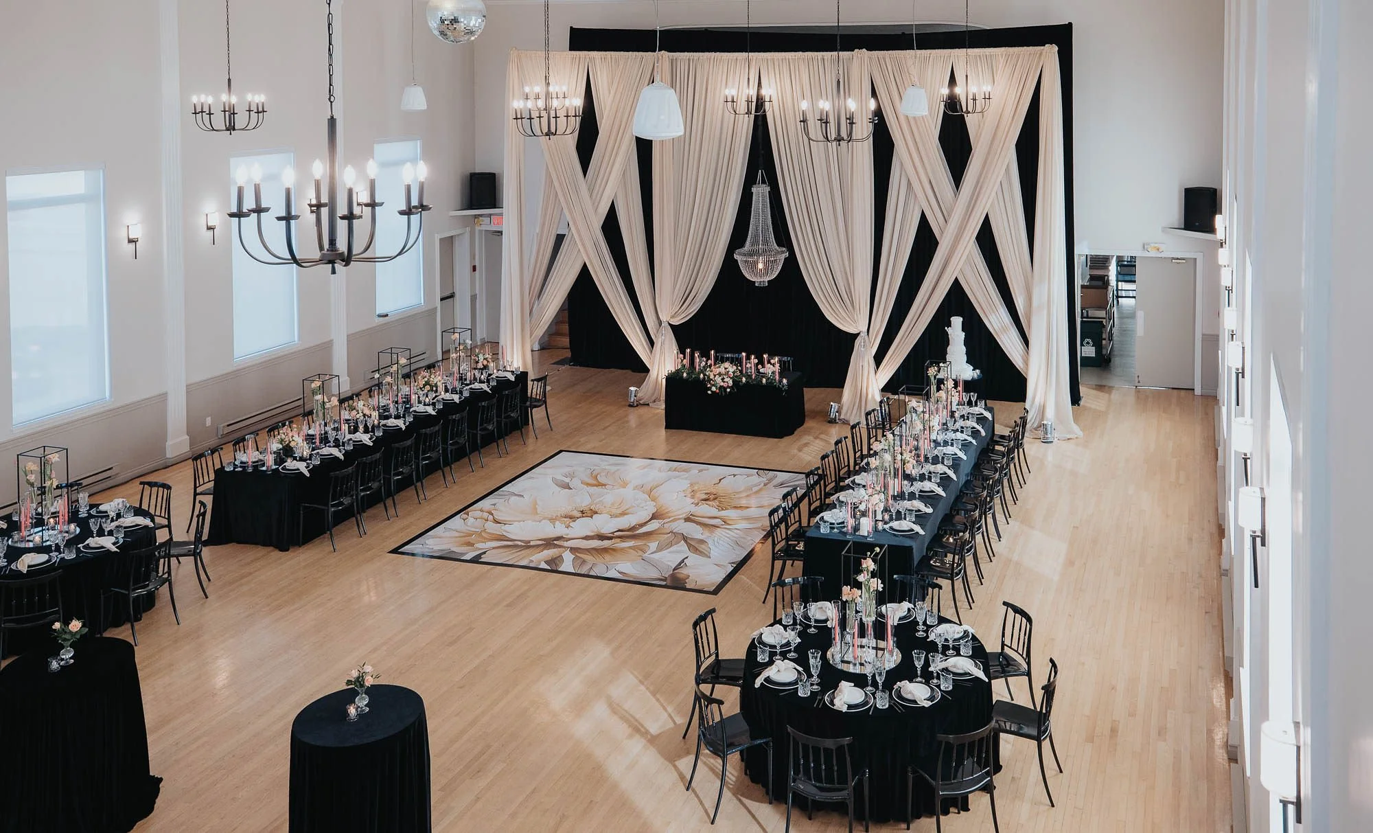 Lavish Ballroom decorated for a wedding or special event, featuring black tablecloths, floral centerpieces, chandeliers, a large floral rug centered in the room, and a black and cream draped backdrop with crystal chandelier at the front.