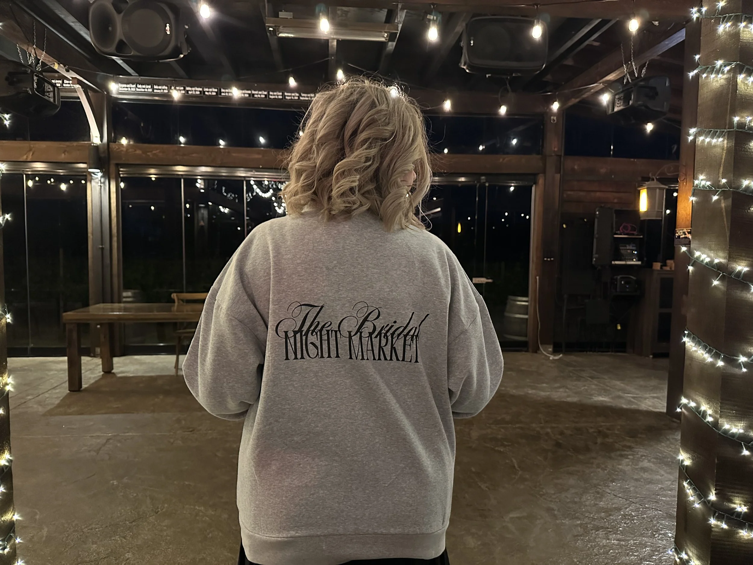 A woman with blonde, curly hair standing in an indoor space decorated with string lights, facing away from the camera. She is wearing a light gray sweatshirt that has 'The Bride Night Market' written on the back in black cursive and uppercase letters