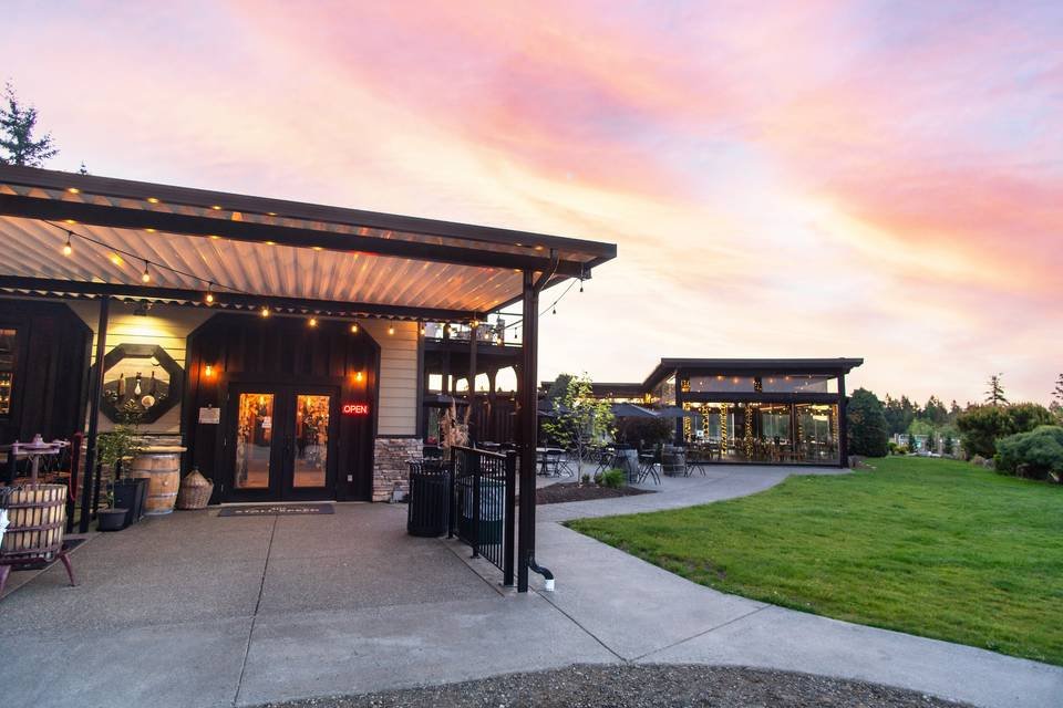Outdoor restaurant or café with glass-enclosed seating areas, string lights, and a grassy lawn under a pink and purple sunset sky at 40 Knots Winery in Comox Valley.