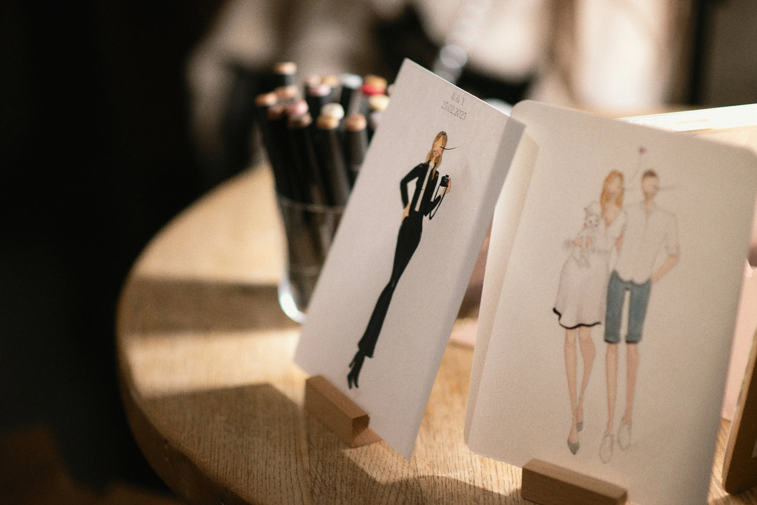 Illustrated wedding invitation cards on a wooden surface, with a container of colored pencils in the background.
