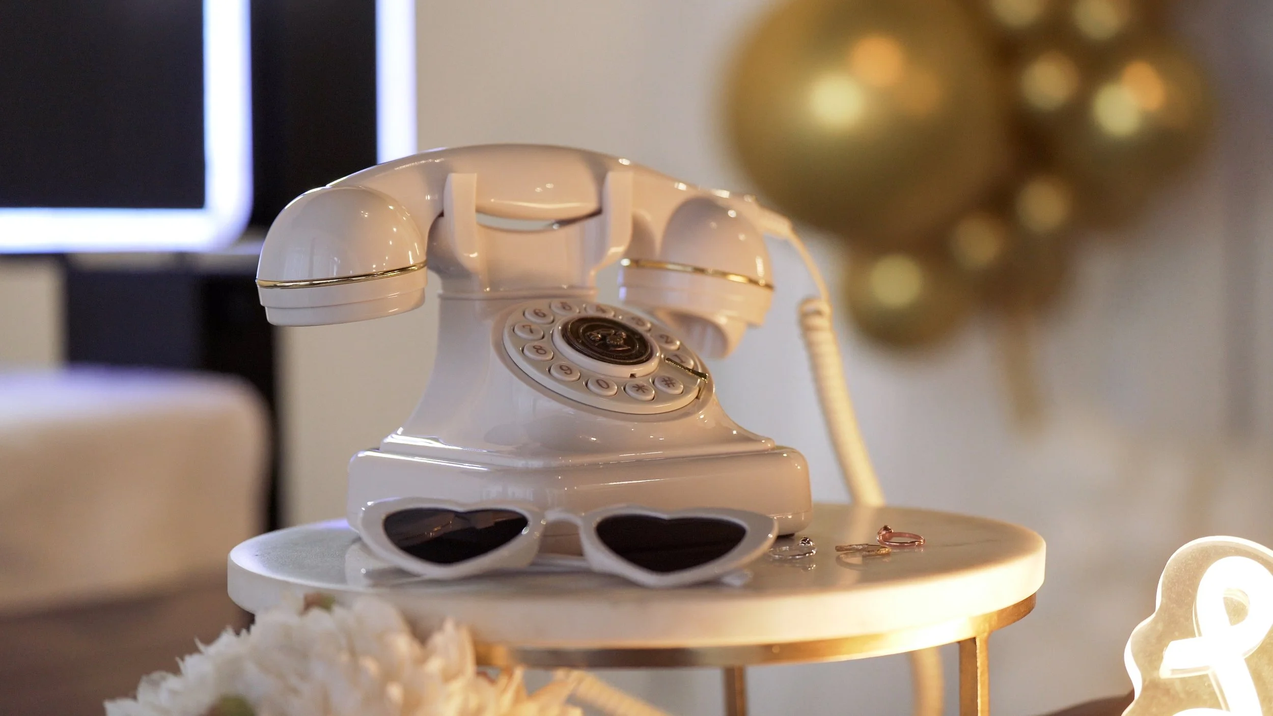 A vintage white rotary telephone decorated with heart-shaped sunglasses placed on top, sitting on a small white and gold side table. In the background, there are blurred gold ornaments and a decorative light. Mirror Mirror Photobooth
