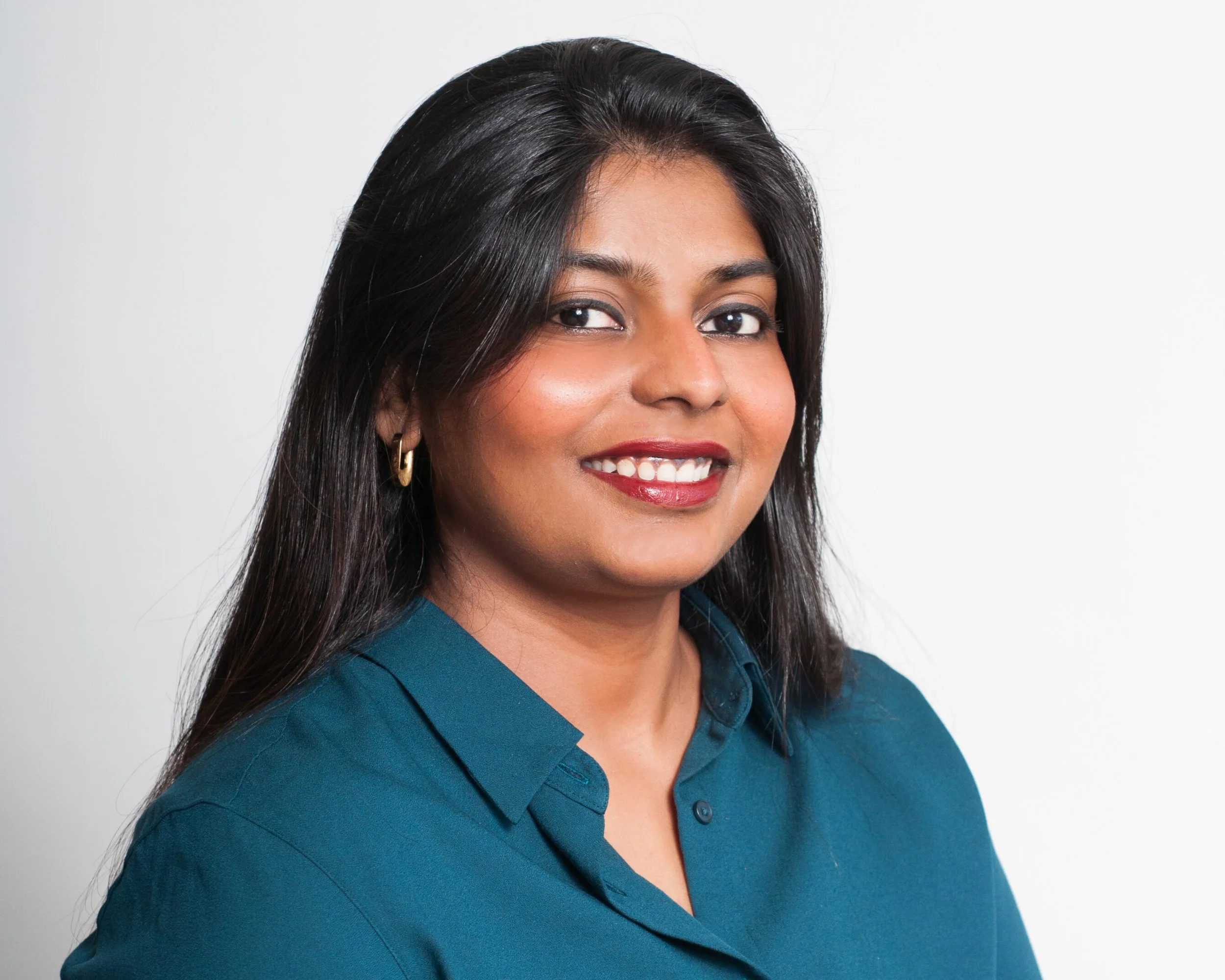 Portrait of a woman with long black hair, wearing gold hoop earrings and a teal button-down shirt, smiling against a white background, Sherin Samuel