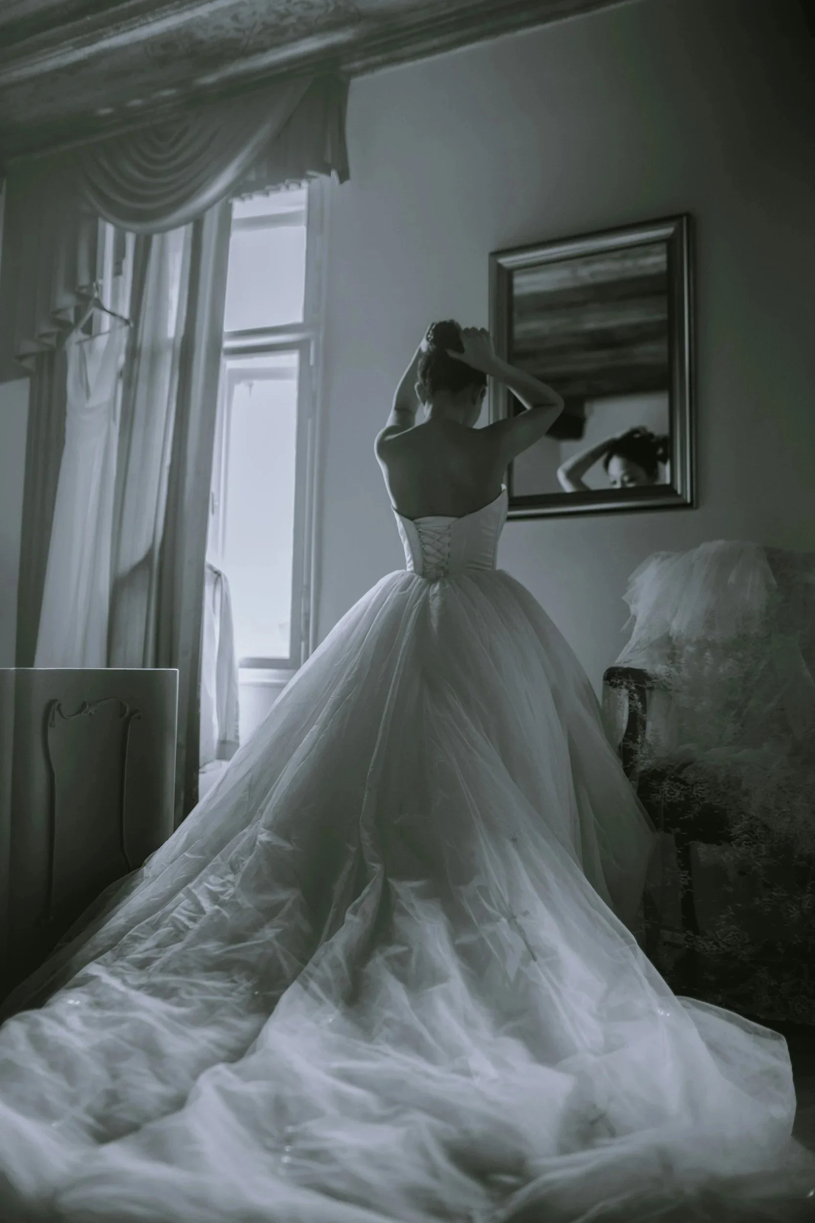 A woman in a wedding dress stands in front of a mirror, adjusting her hair in a softly lit room with large windows and elegant curtains.