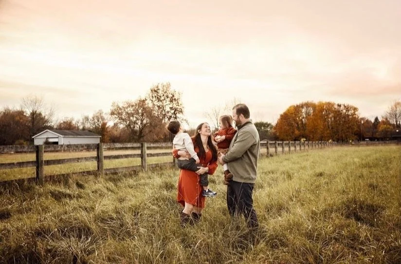 A family of four standing in a grassy field with a wooden fence, trees, and a house in the background during sunset.