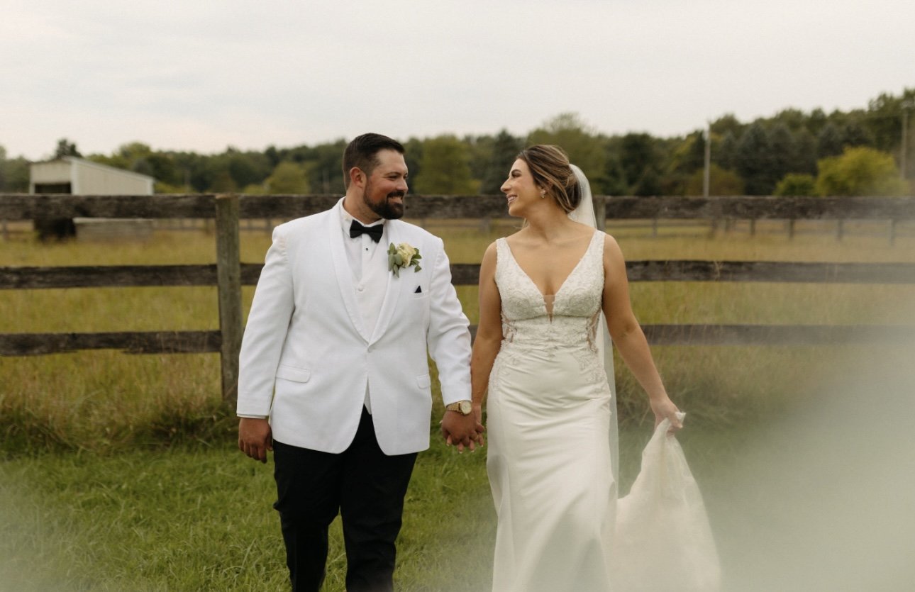 A newlywed couple walking hand in hand outdoors in a grassy field, with a wooden fence and trees in the background on a cloudy day.