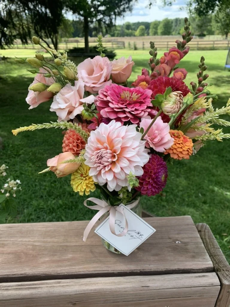 A colorful bouquet of pink, peach, yellow, orange, and purple flowers in a glass jar with a white ribbon and a handwritten note, set outdoors on a wooden surface with a green grassy field and trees in the background.