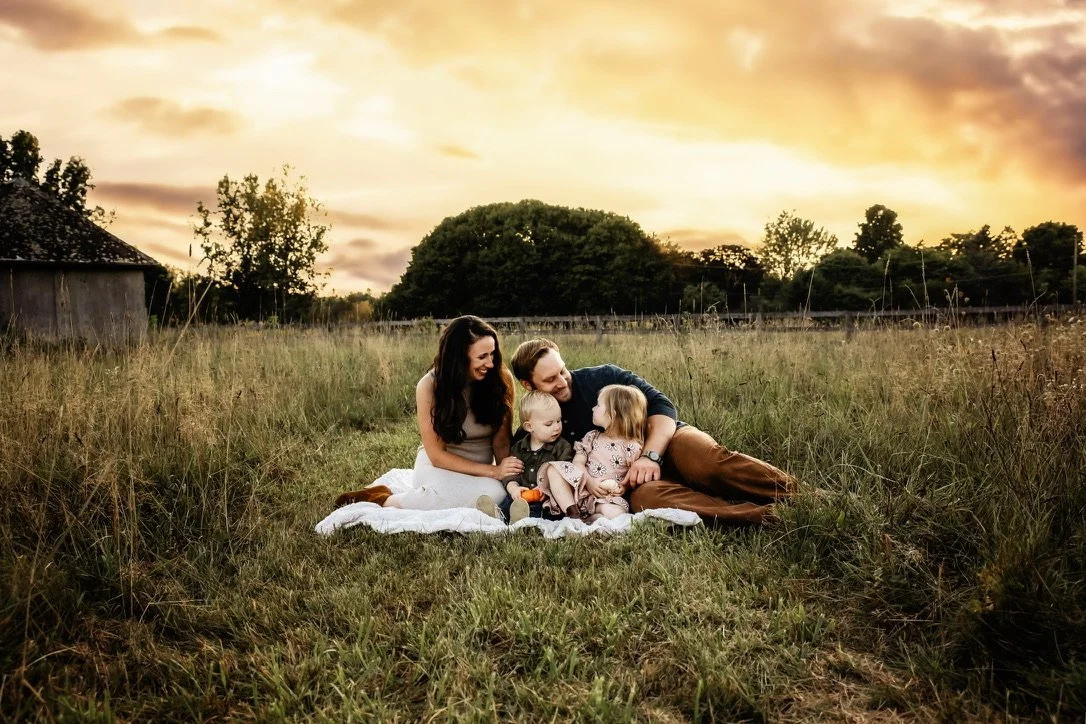Family sitting on a blanket in a grassy field at sunset, including two adults and two young children, with trees and an old barn in the background.