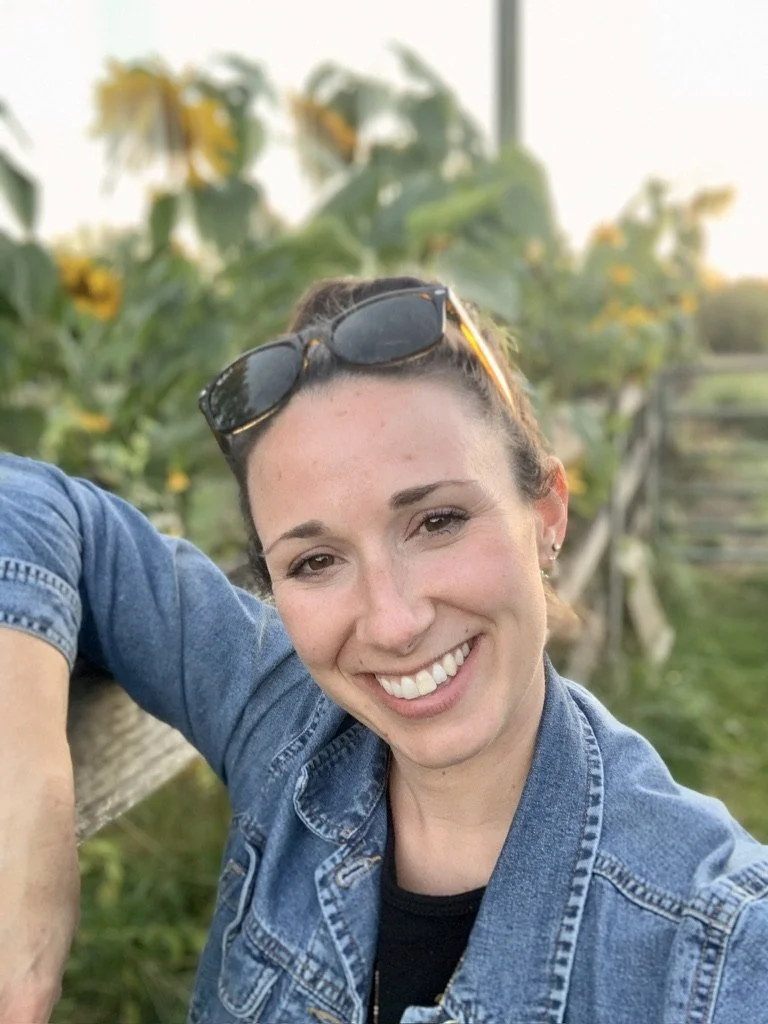 A woman outdoors smiling, with sunglasses on her head, wearing a denim jacket, and sunflowers in the background.