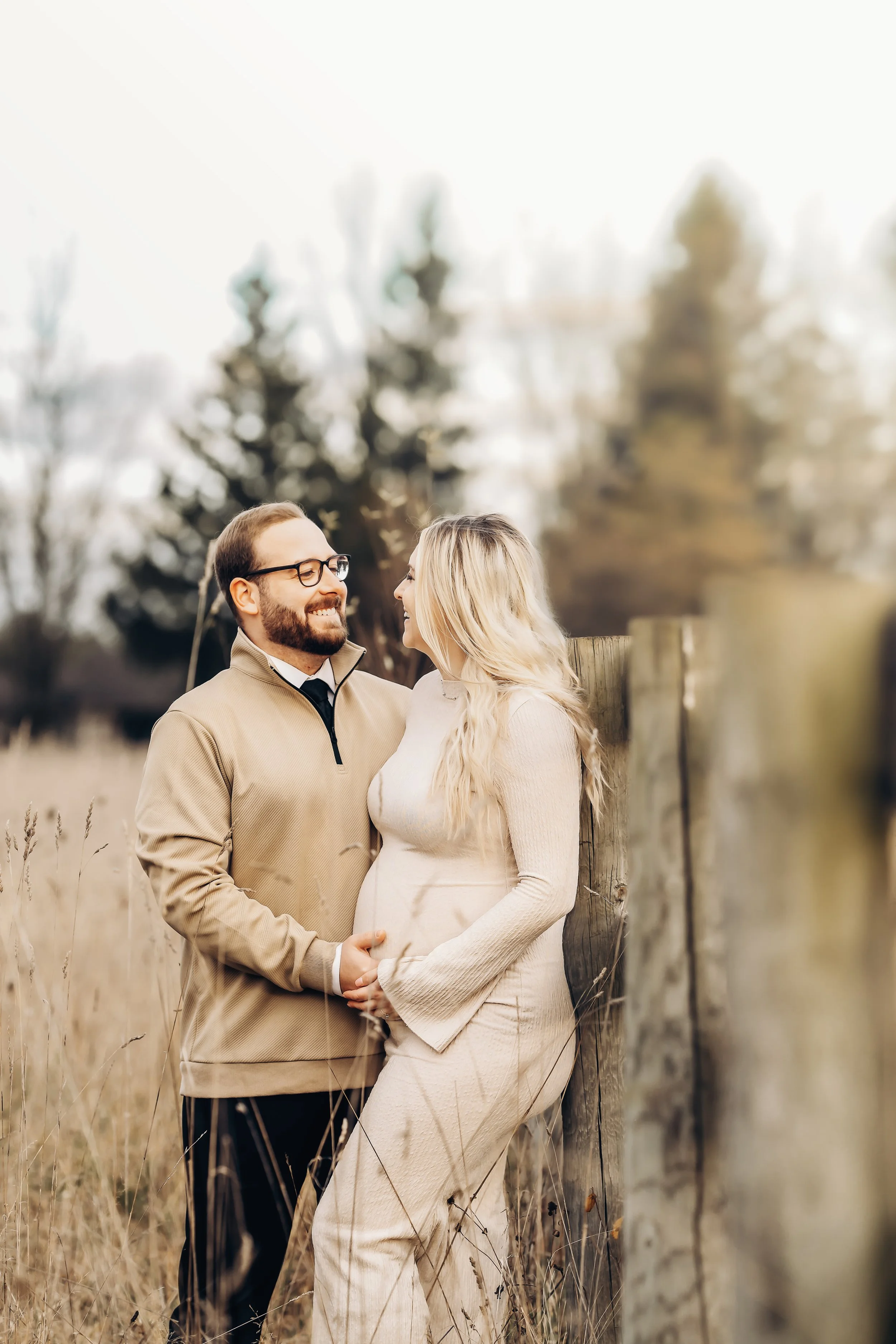 A happy couple standing outdoors in a field, facing each other and smiling. The woman is pregnant and gently touching her belly, while the man is holding her hand. They are leaning against a wooden fence with trees in the background, on an overcast day.