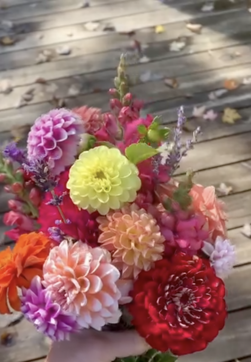 Close-up of a colorful bouquet of flowers, including pink, yellow, orange, purple, and red blooms, held in a person's hand over a wooden surface with scattered autumn leaves.