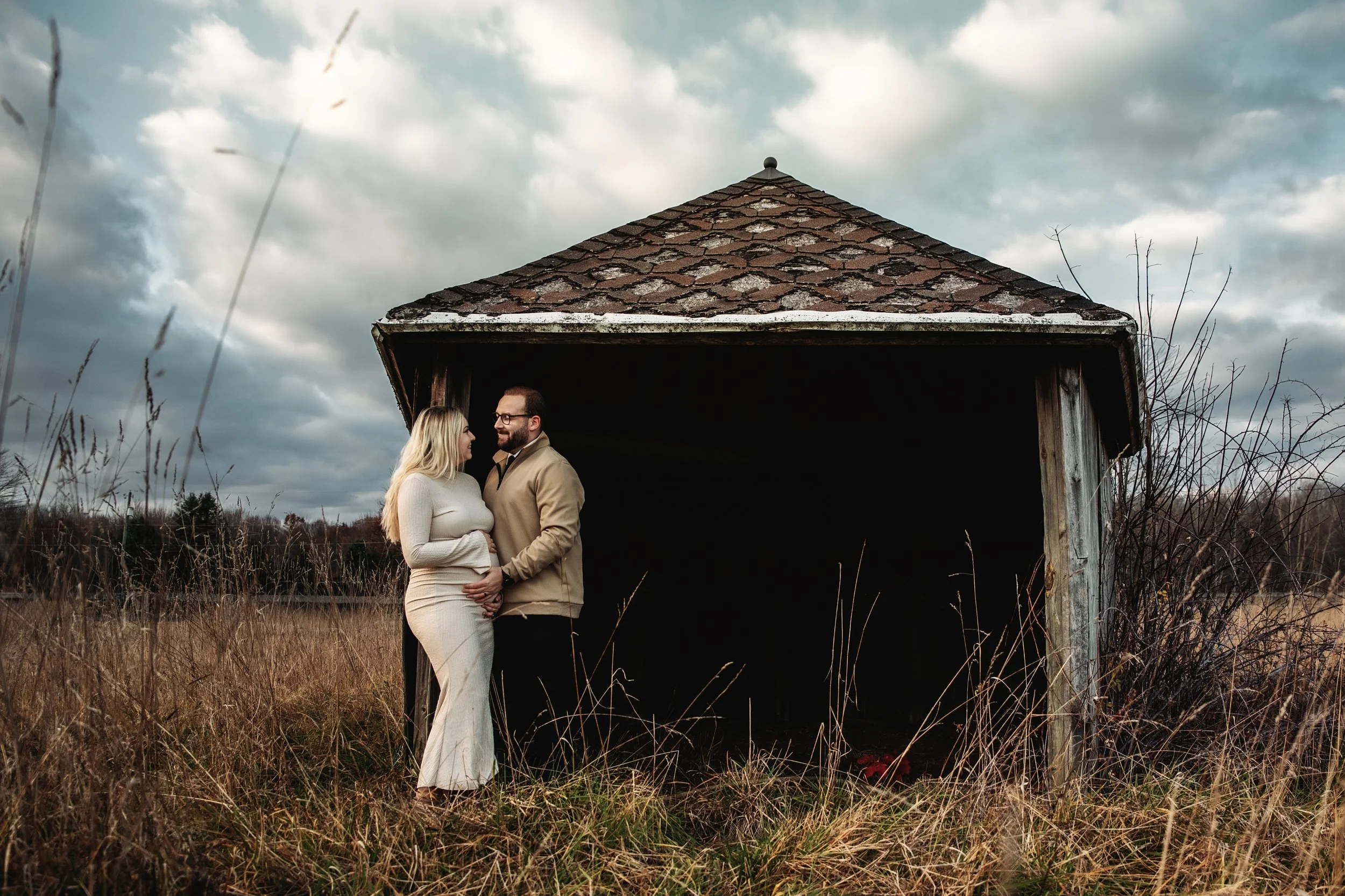 A couple standing close together in a field of tall grass in front of an old, abandoned wooden shelter with a roof, holding hands and looking at each other, with a cloudy sky overhead.