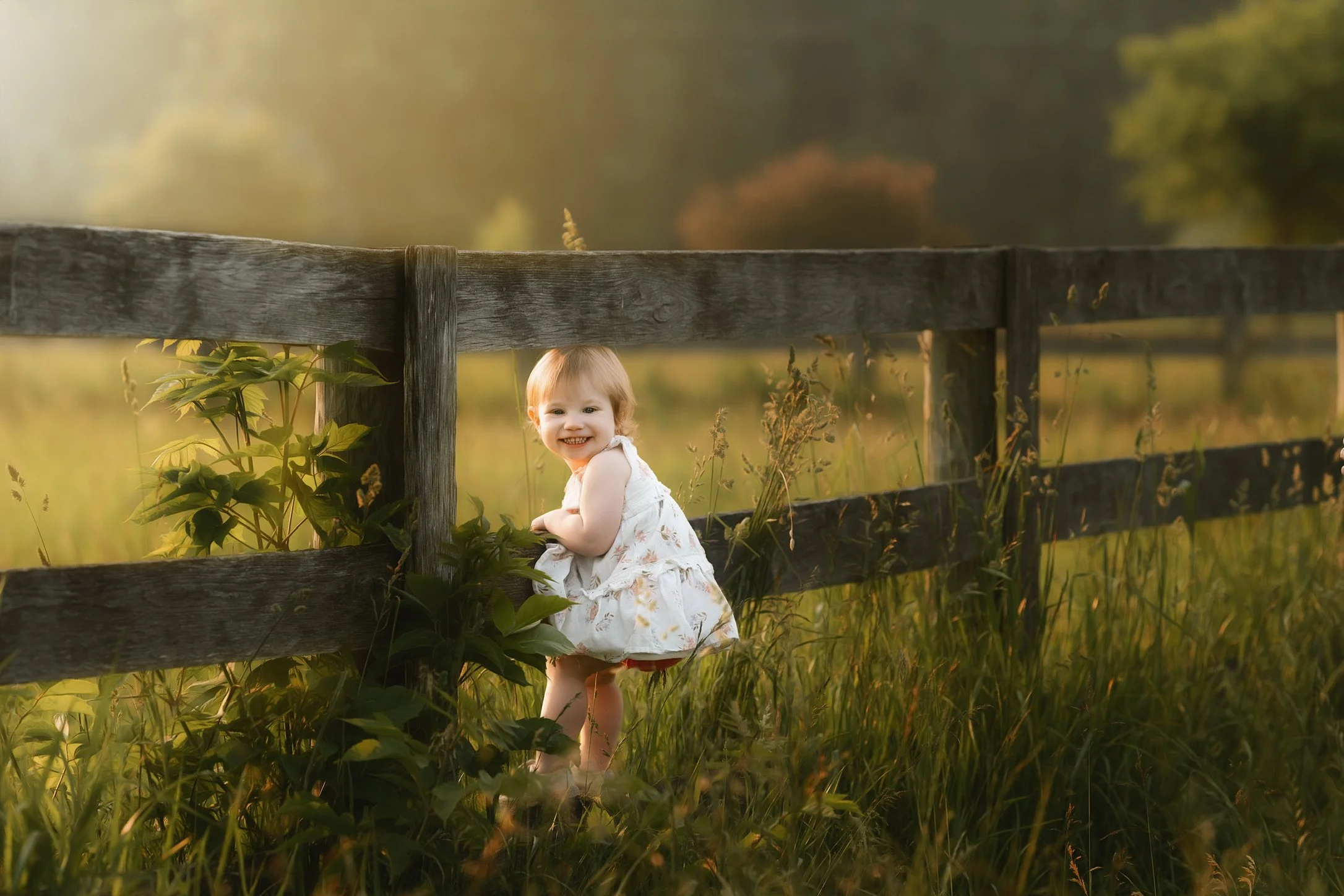 A young girl standing next to a rustic wooden fence in a grassy field during sunset, smiling at the camera.