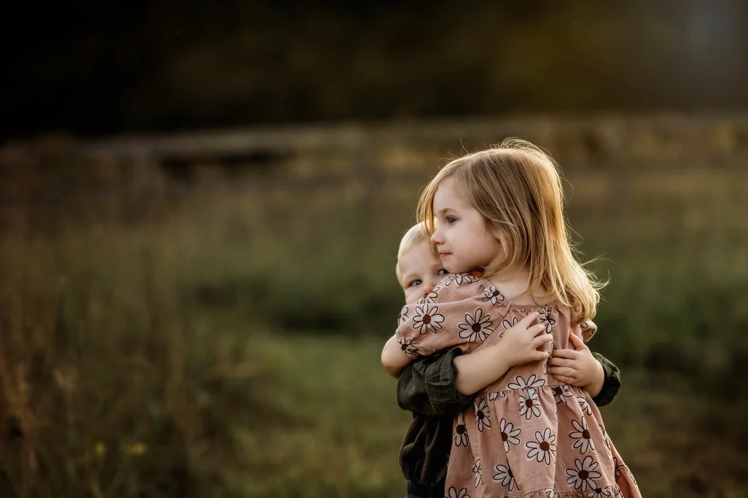 Two young children, a girl with reddish hair and a boy with blonde hair, share a warm hug outdoors by a body of water in a natural setting during sunset.