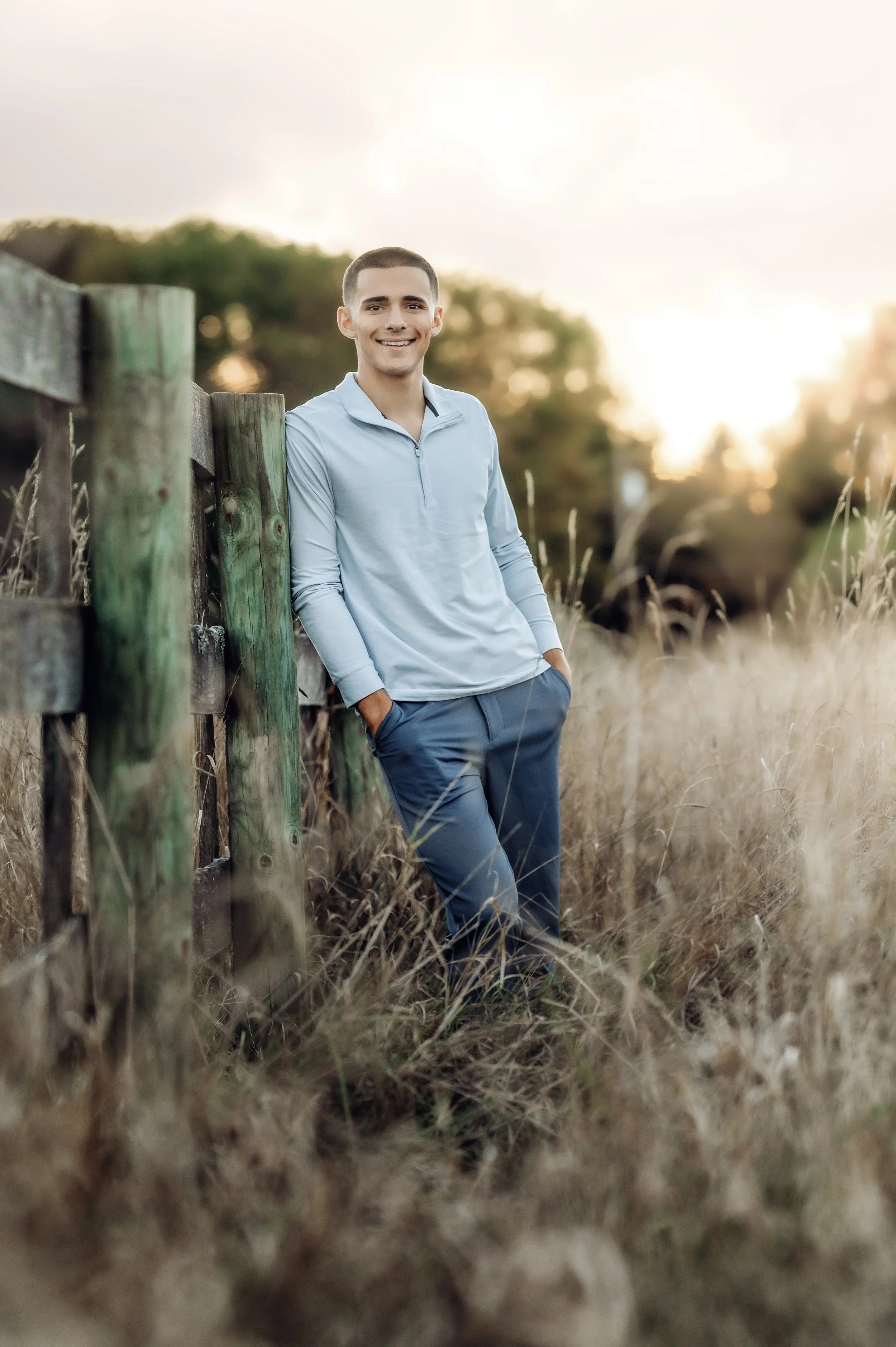 A young man in a light blue long-sleeve shirt and blue pants standing outdoors by a wooden fence during sunset, smiling and looking at the camera.