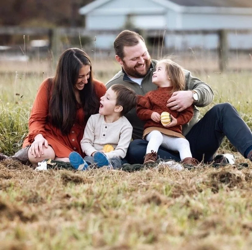 Family of four sitting on grass outdoors, smiling and laughing together. The mother, father, boy, and girl are enjoying a happy moment, holding small figs or apples.