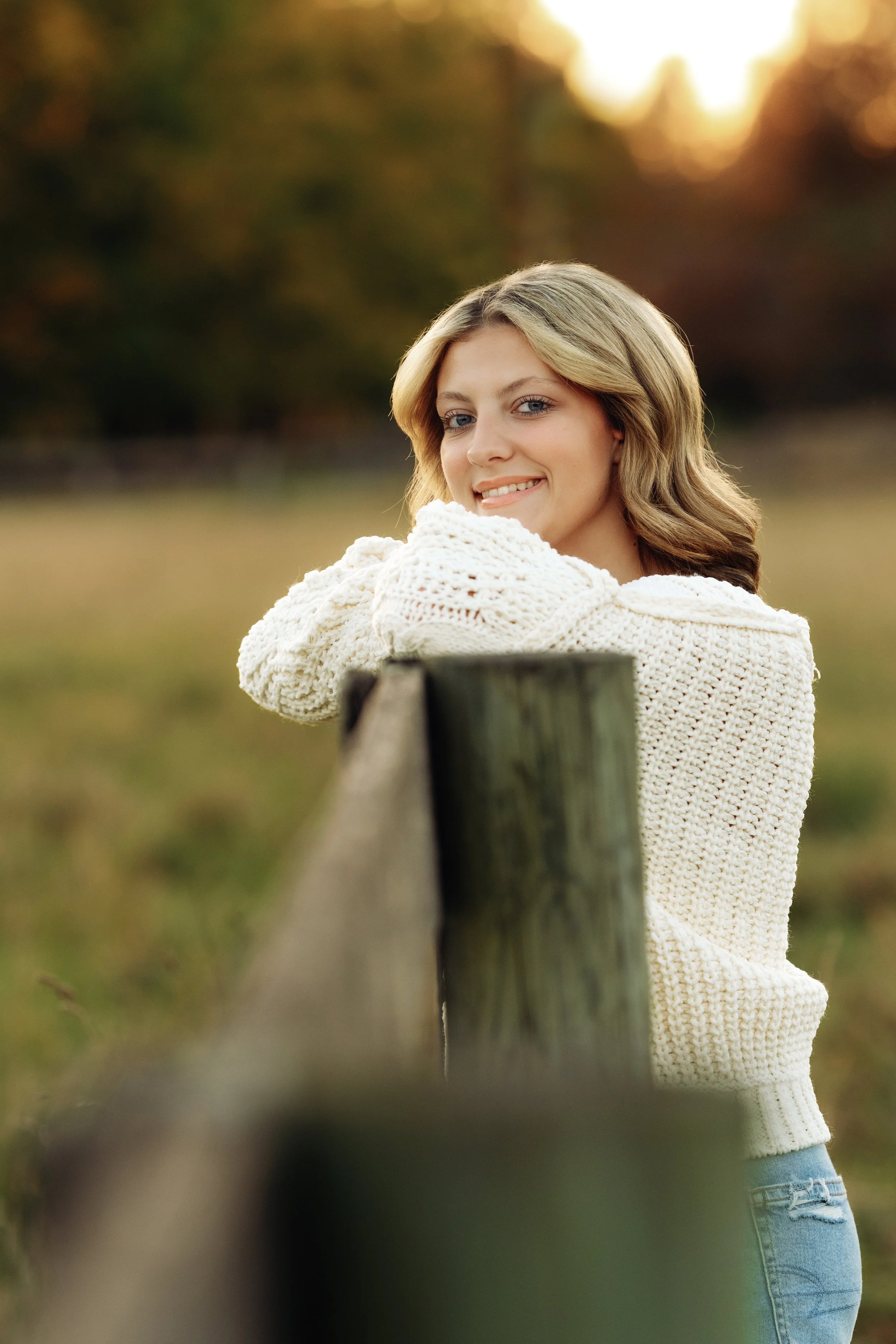 A young woman with blonde hair wearing a white knitted sweater and light blue jeans leaning on a wooden fence outdoors during fall.