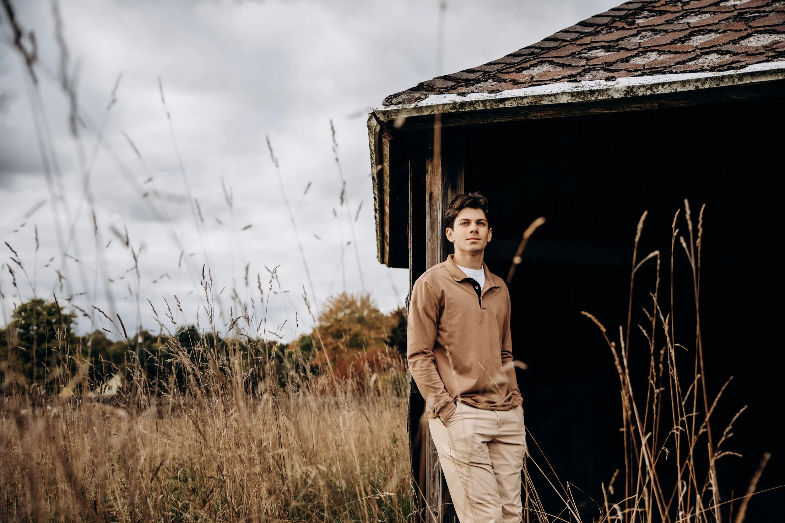 A young man with dark hair standing outdoors in front of an old wooden structure, with tall grass and cloudy sky in the background.