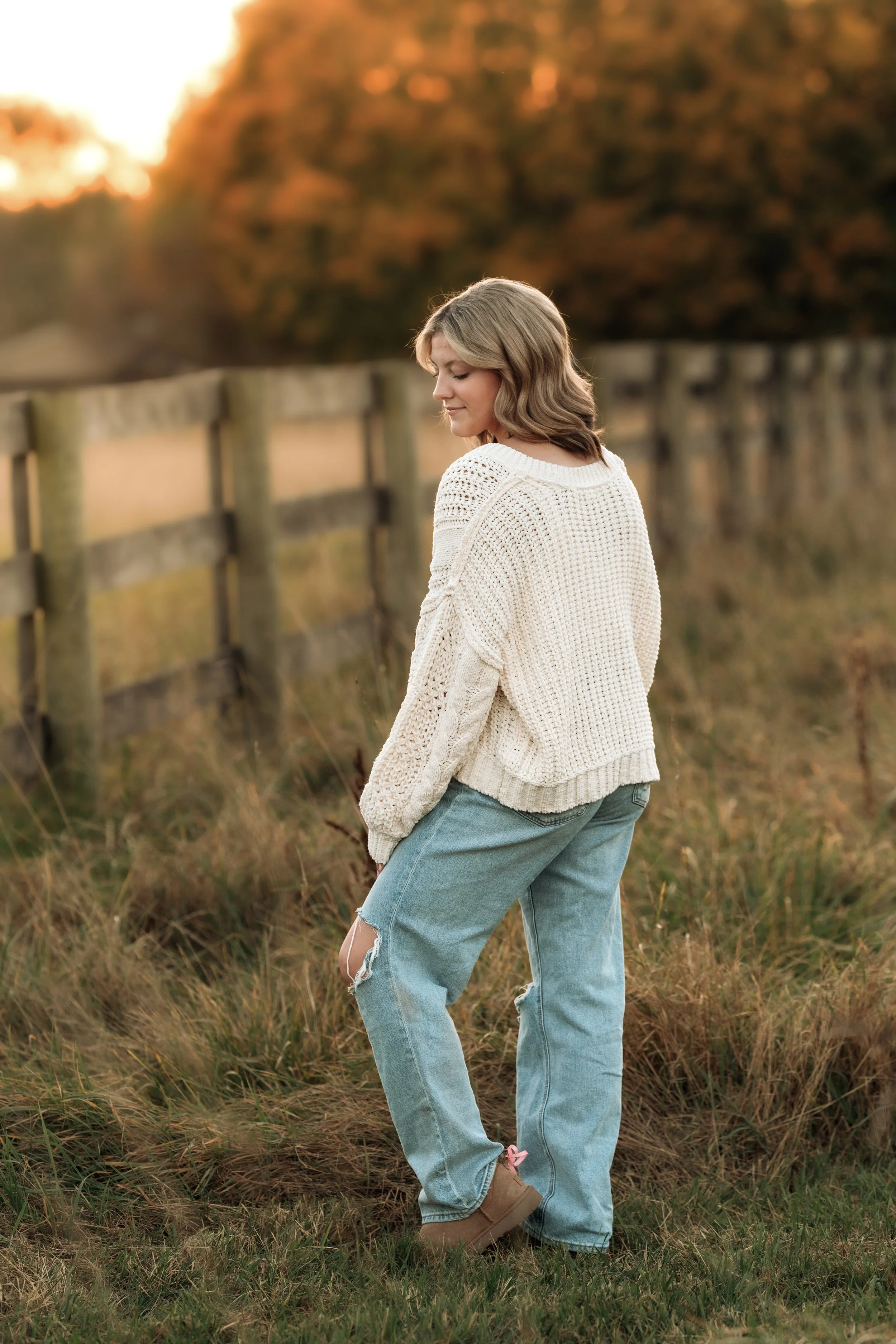 A woman with short blond hair wearing a cream knitted sweater, ripped jeans, and tan boots standing outdoors at sunset with a wooden fence and autumn trees in the background.
