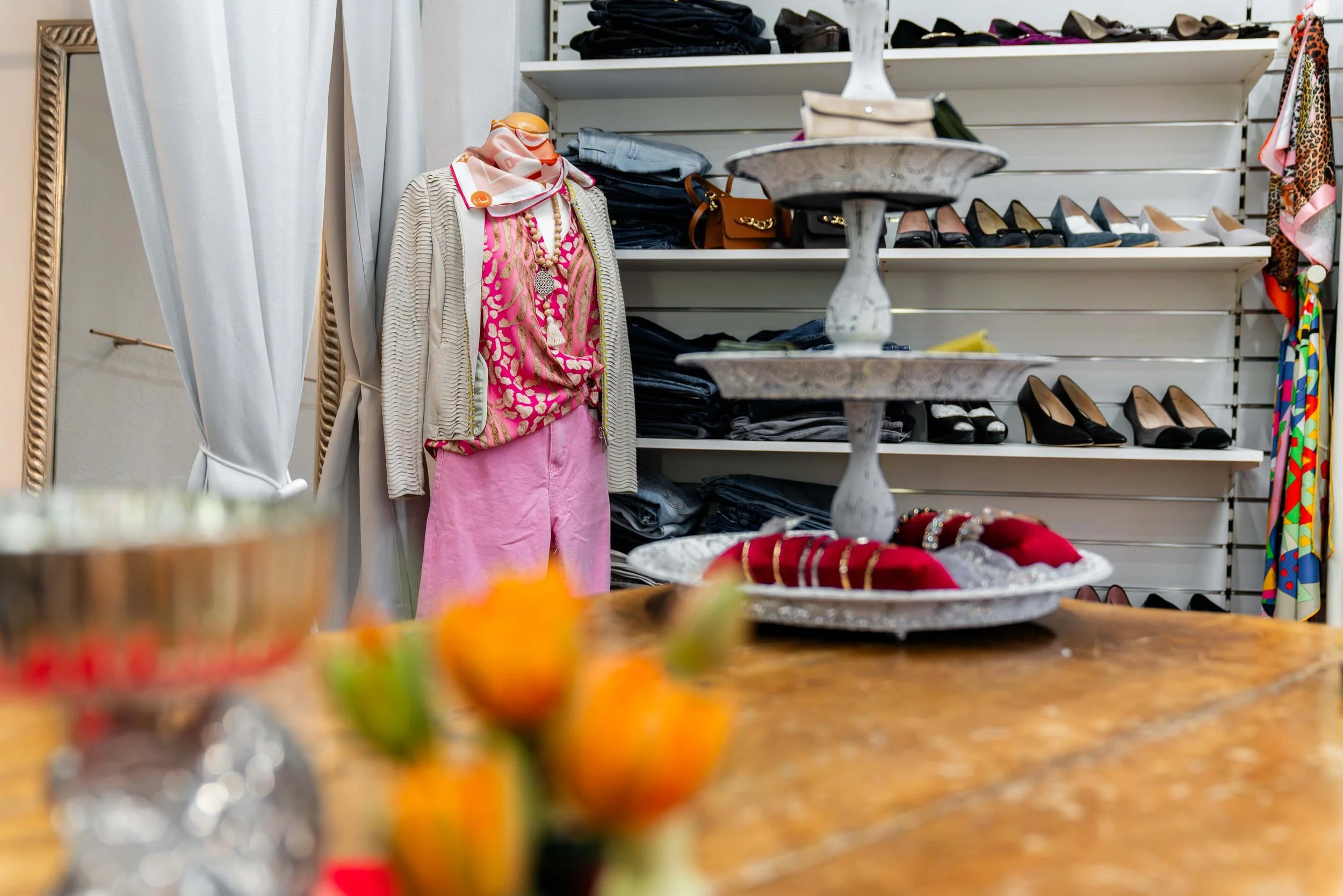 Hintergrund eines Kleiderladens mit Schaufenster, darin eine Puppe mit pinker Hose, rosa Top, beigem Strickjacke und Schal, Regale mit hohen Absätzen, auf einem Tisch Schmuck und Schuhe, im Vordergrund eine Vase mit Tulpen.