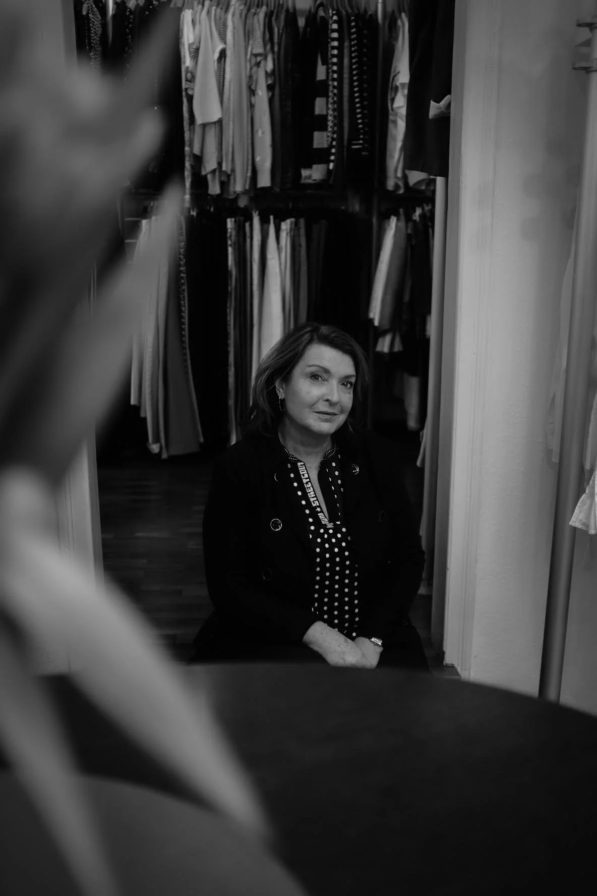Inhaberin von Der Besondere Laden sitzt in der Boutique vor einem Kleiderschrank mit Designer-Second-Hand-Kleidung, Schwarz-Weiß-Aufnahme.