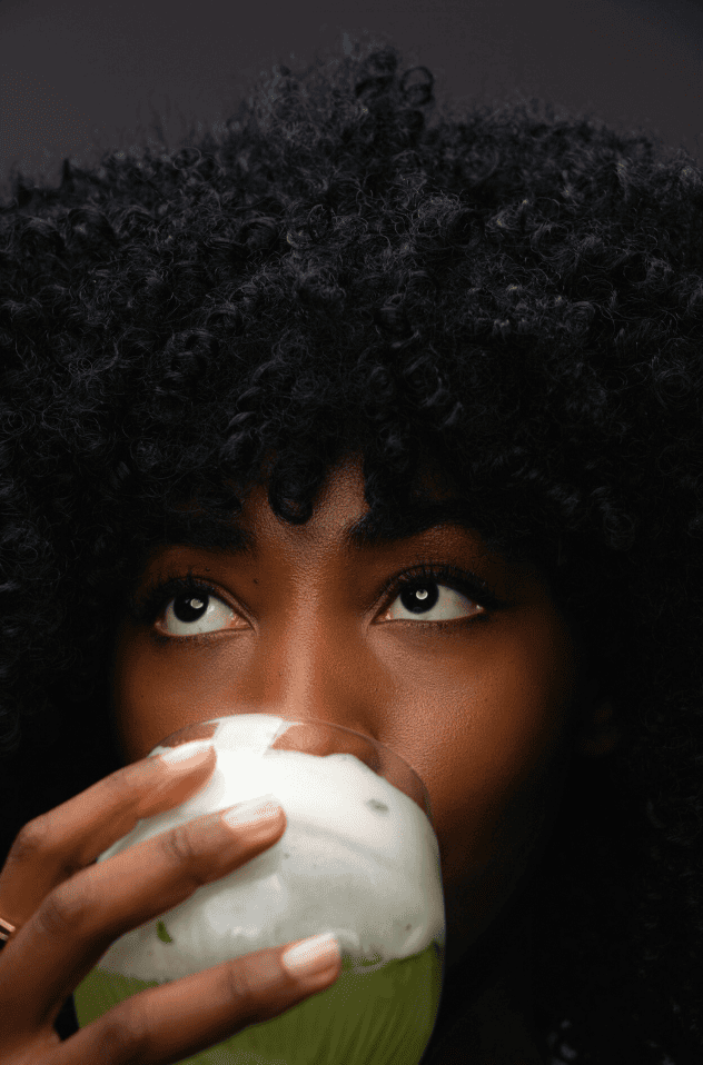 A woman with dark curly hair holding and drinking from a green and white coconut.