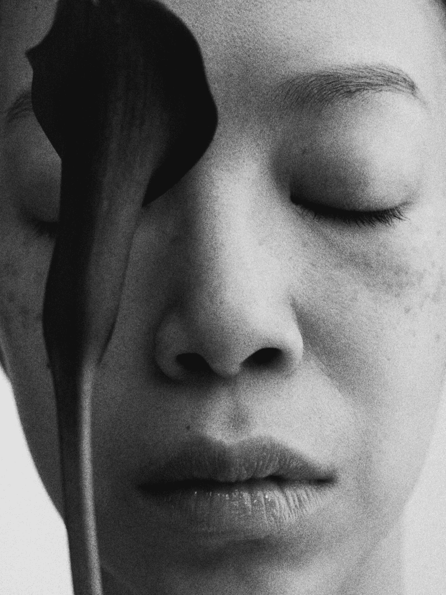 Close-up black and white photograph of a person's face with closed eyes, holding a dark flower near their cheek.