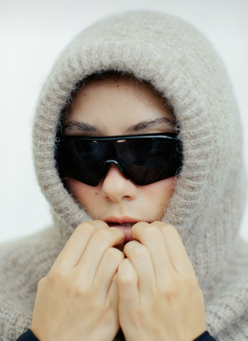 Person wearing a beige hooded sweater, large black sunglasses, and holding hands near mouth with a contemplative expression.